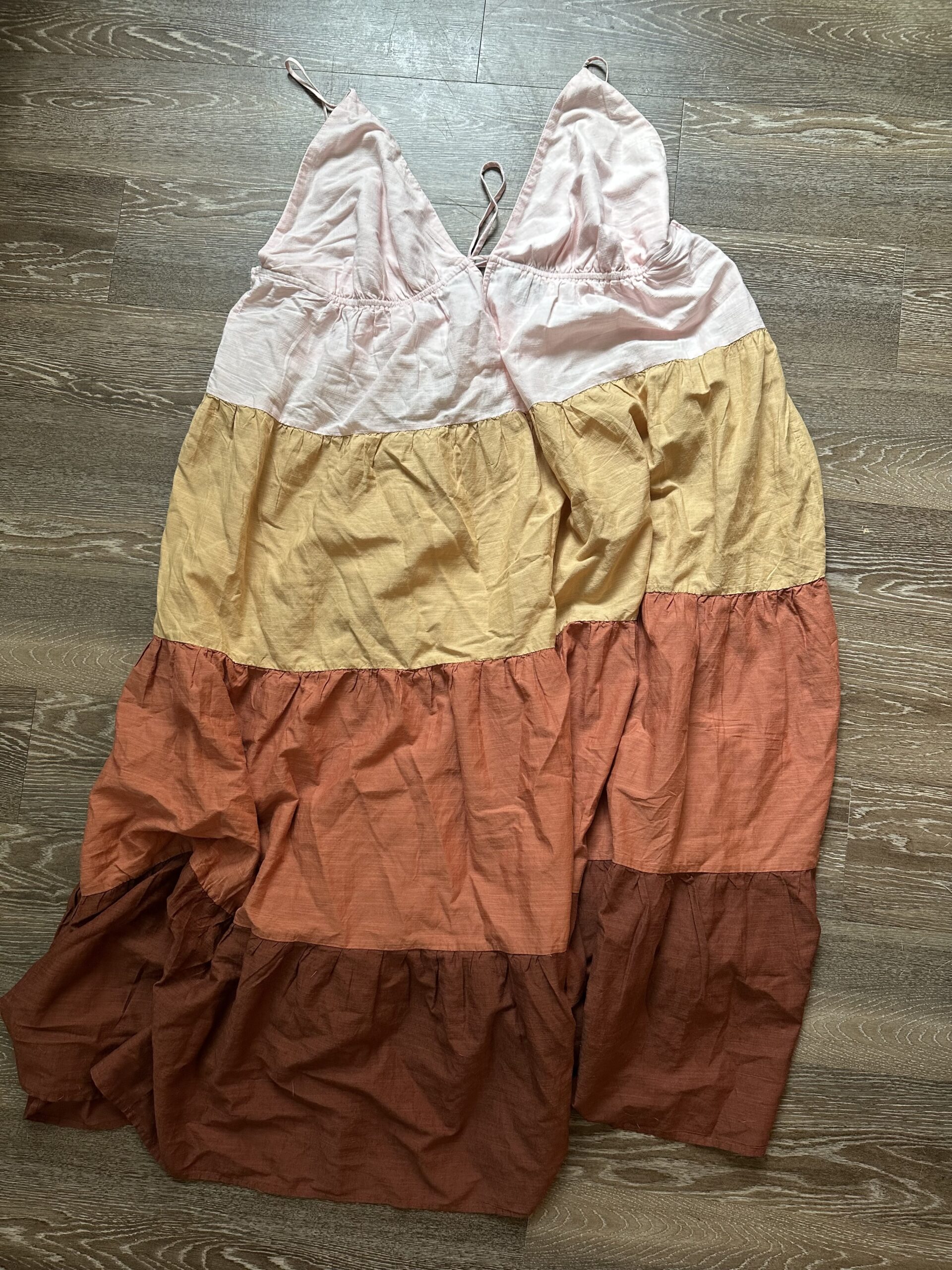 A sleeveless, tiered summer dress with horizontal stripes in shades of pink, beige, yellow, and red, displayed on a wooden floor.
