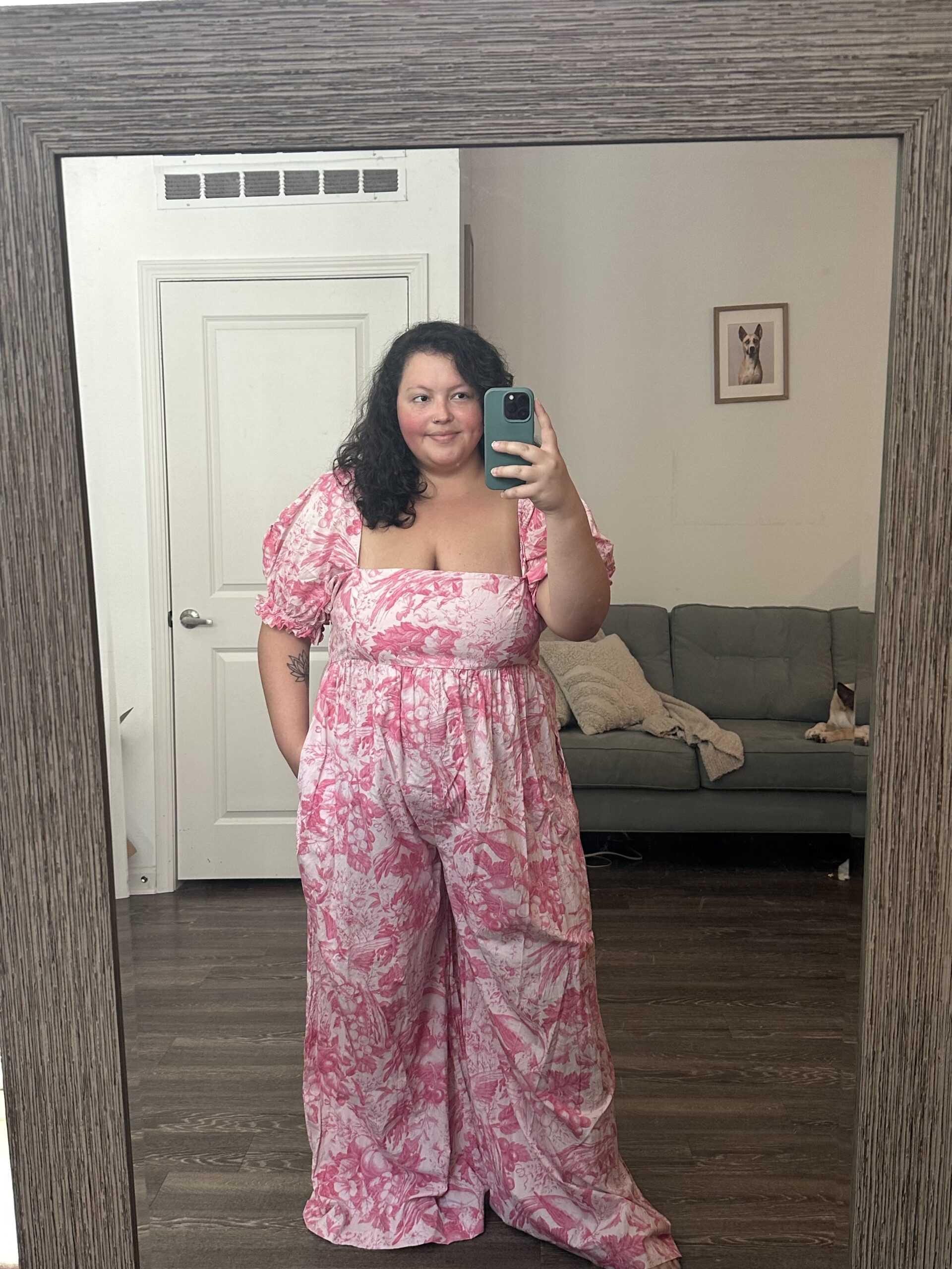 A woman in a pink floral dress takes a selfie in a mirror with a phone, with a living room visible in the background.