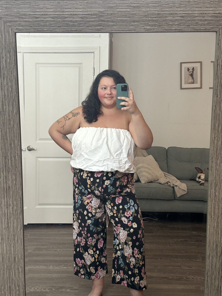 Woman in a white top and floral pants taking a mirror selfie in a room with a couch and framed picture in the background.