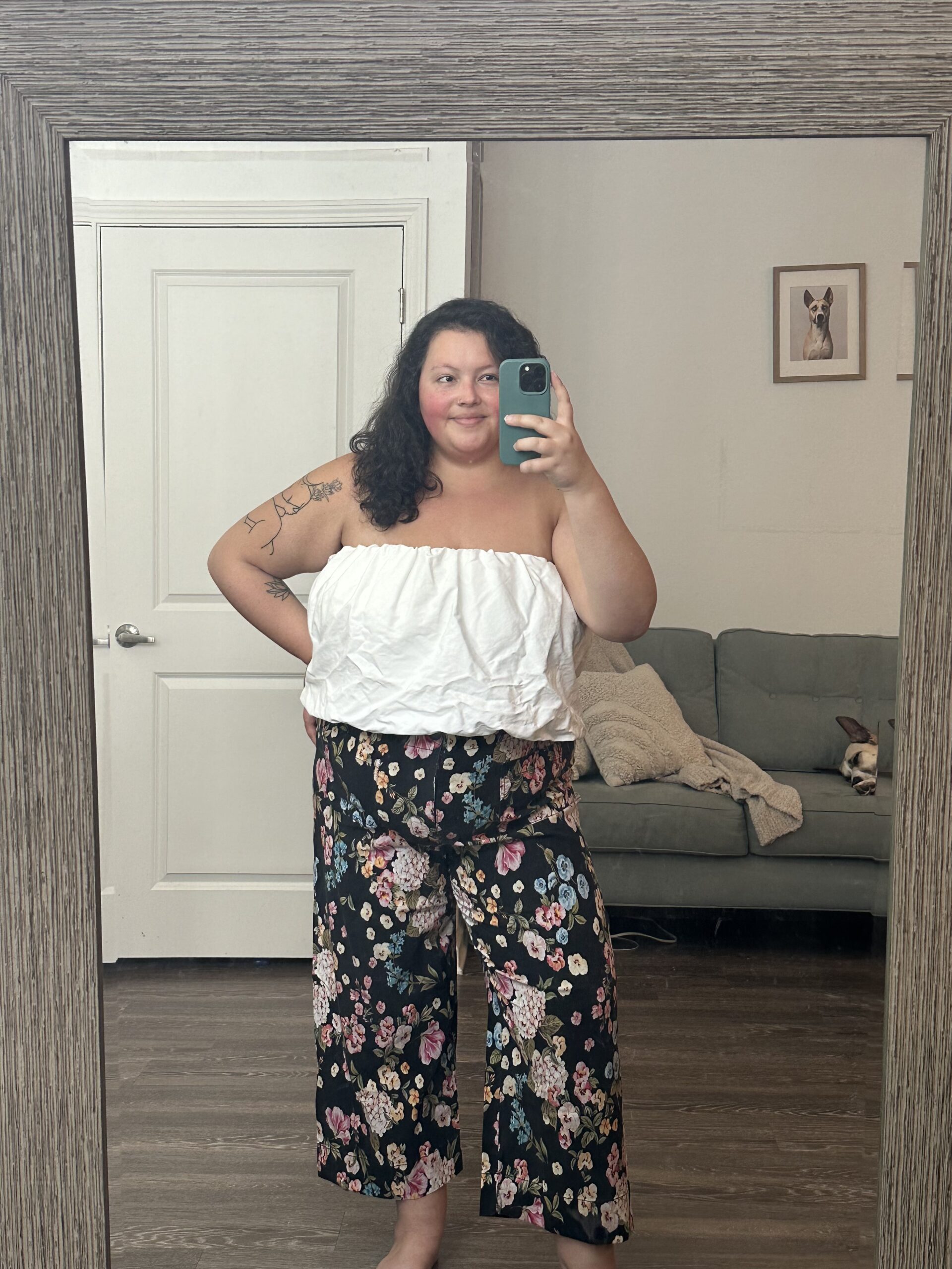 Woman in a white top and floral pants taking a mirror selfie in a room with a couch and framed picture in the background.