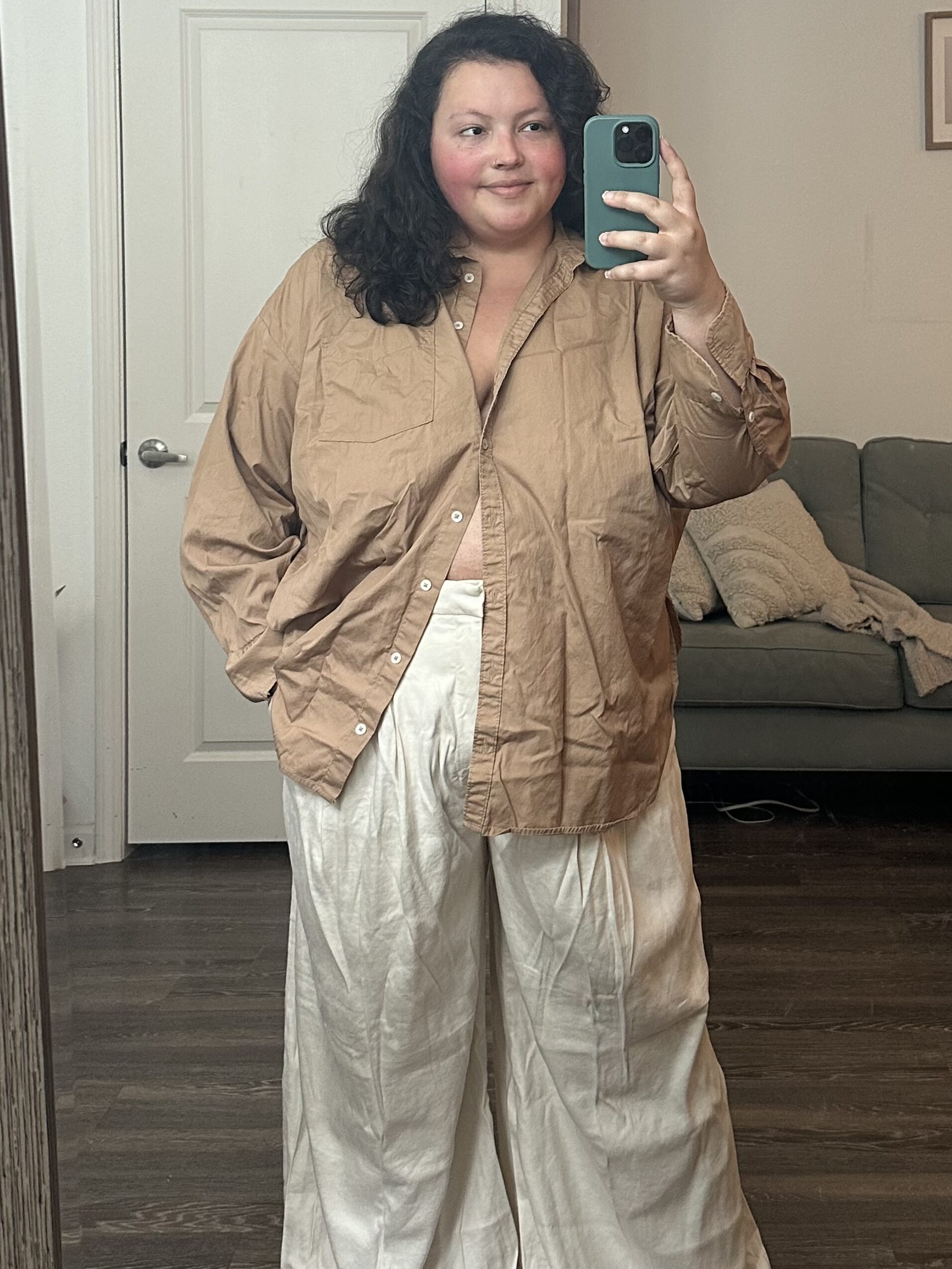 A young woman taking a mirror selfie with a smartphone, wearing a beige jacket and wide cream trousers, a gray sofa and wall art visible in the background.