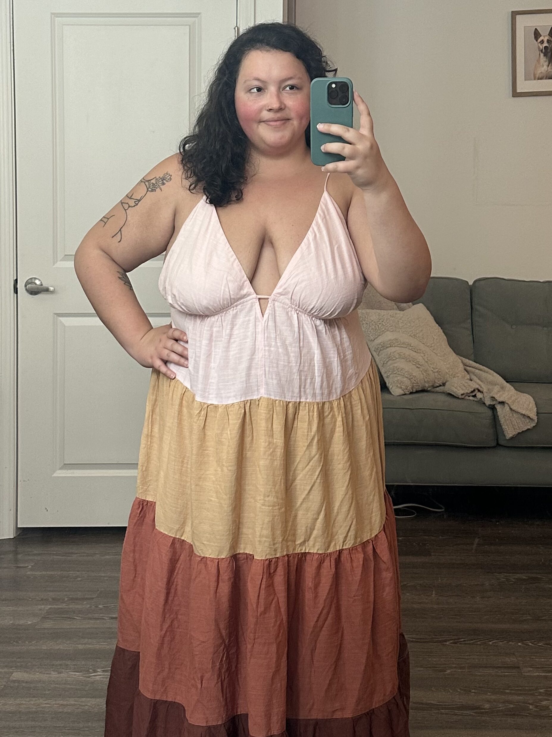 Woman standing in front of a mirror, taking a selfie, wearing a colorful tiered dress, with tattoos visible on her upper arm.