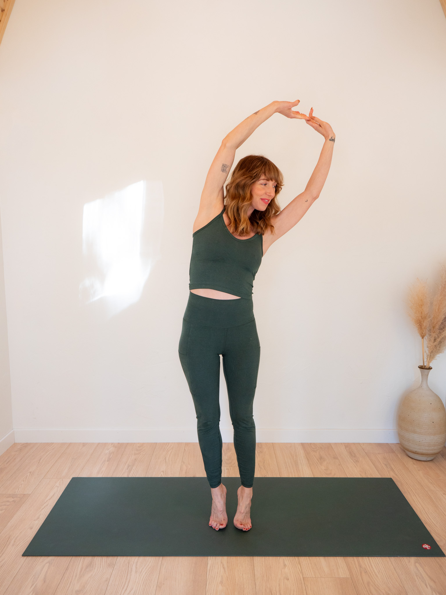 A woman in a green yoga outfit stretches her arms above her head, standing on a yoga mat in a bright, minimalist room.