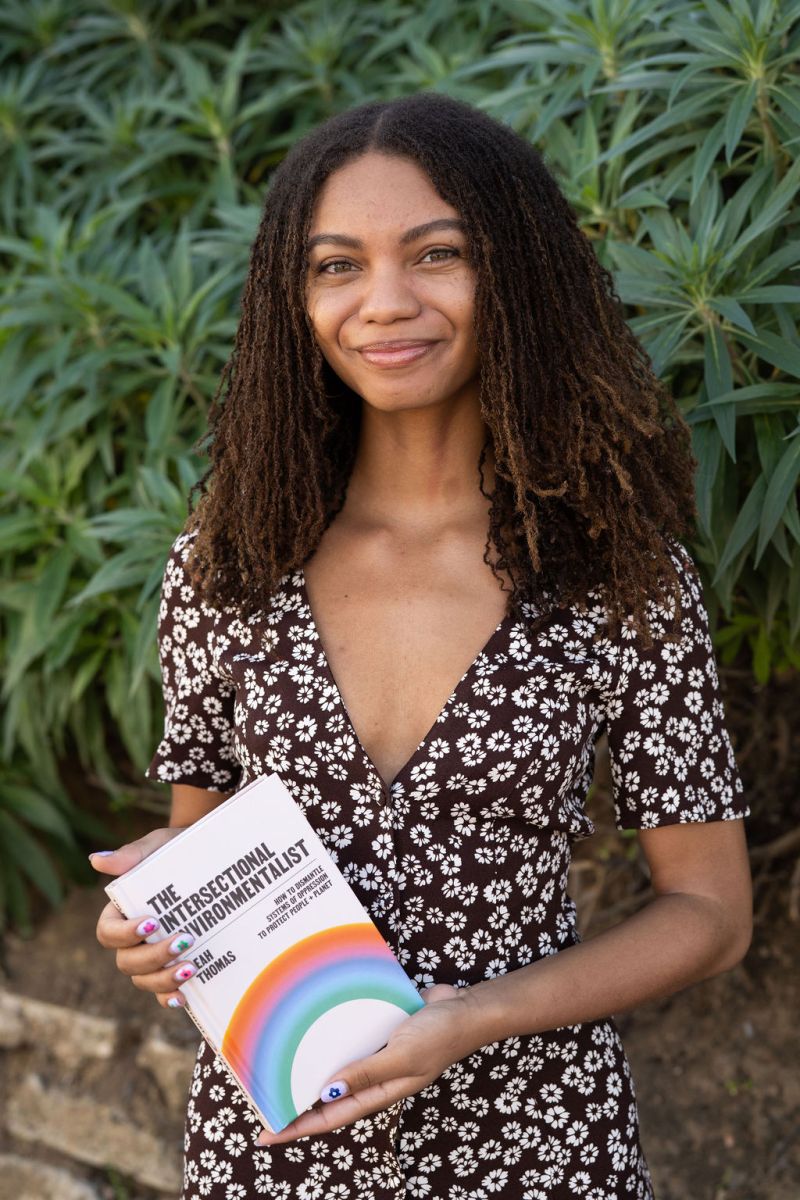 A smiling young woman holding a book titled "the intersectional environmentalist" stands in front of leafy plants.