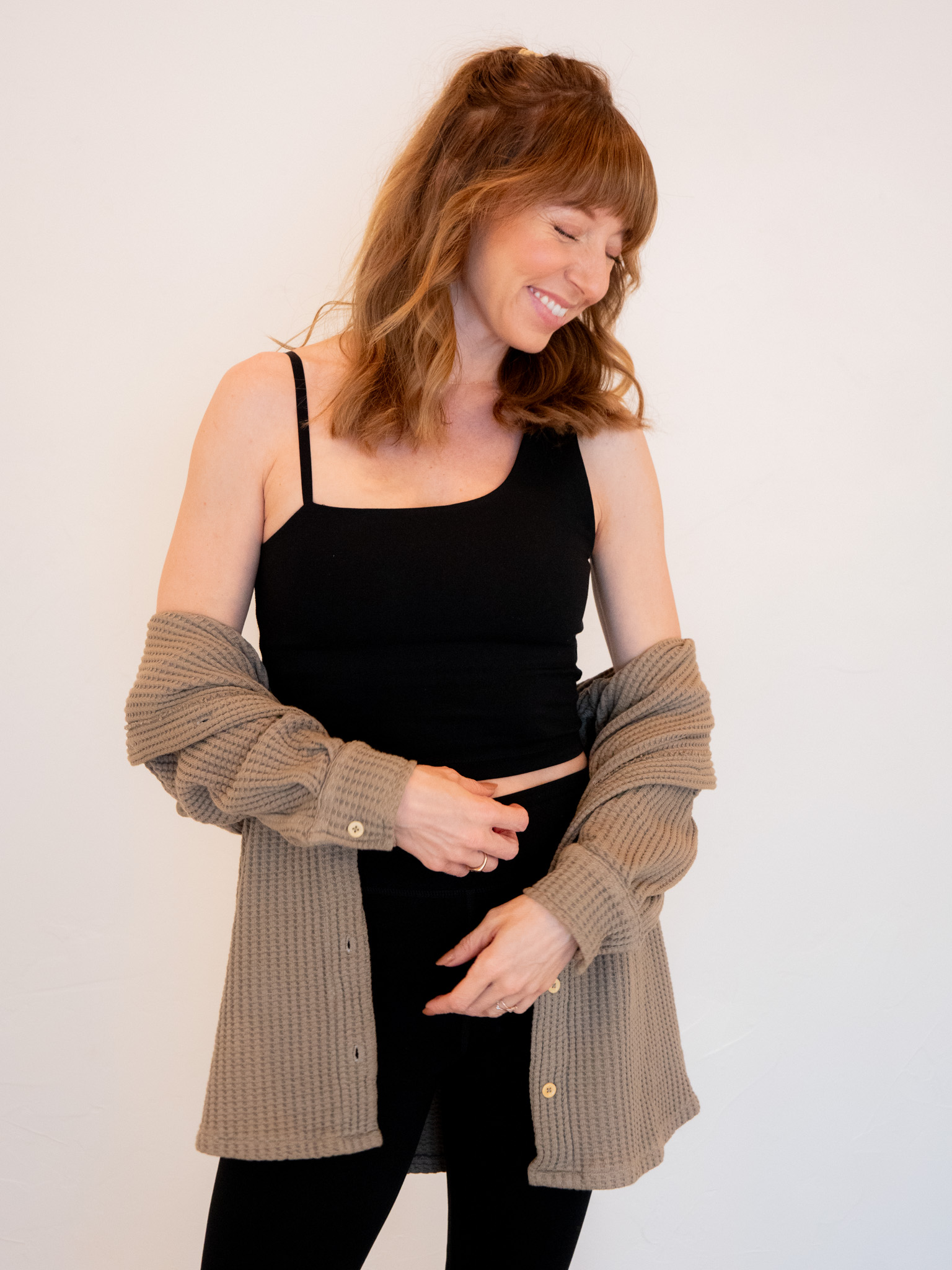 A smiling woman with long brown hair, wearing a black tank top and a beige cardigan, standing against a white background.