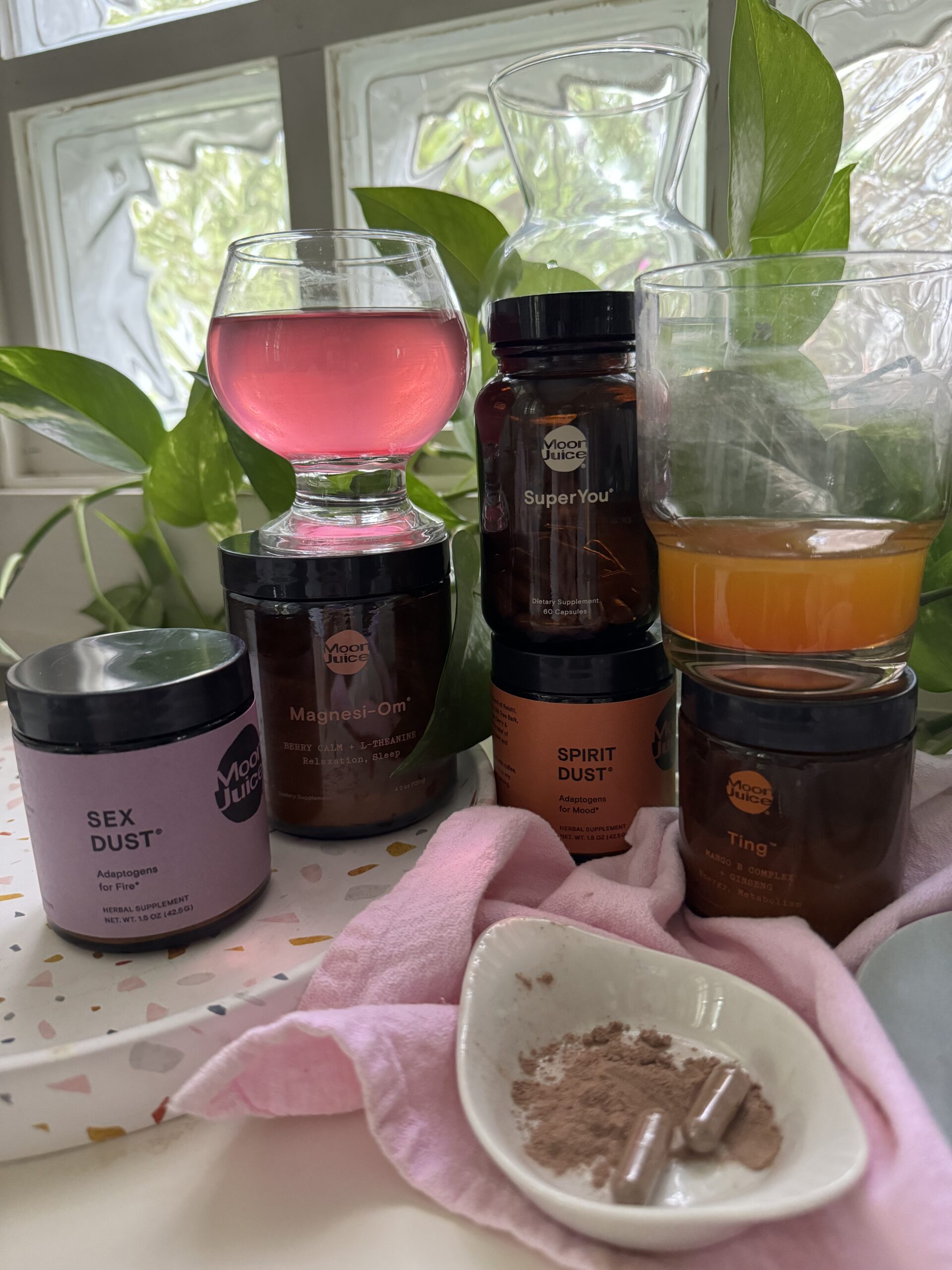 A kitchen counter with a glass of pink liquid, various wellness supplements in jars, and a spoonful of powder on a pink cloth.