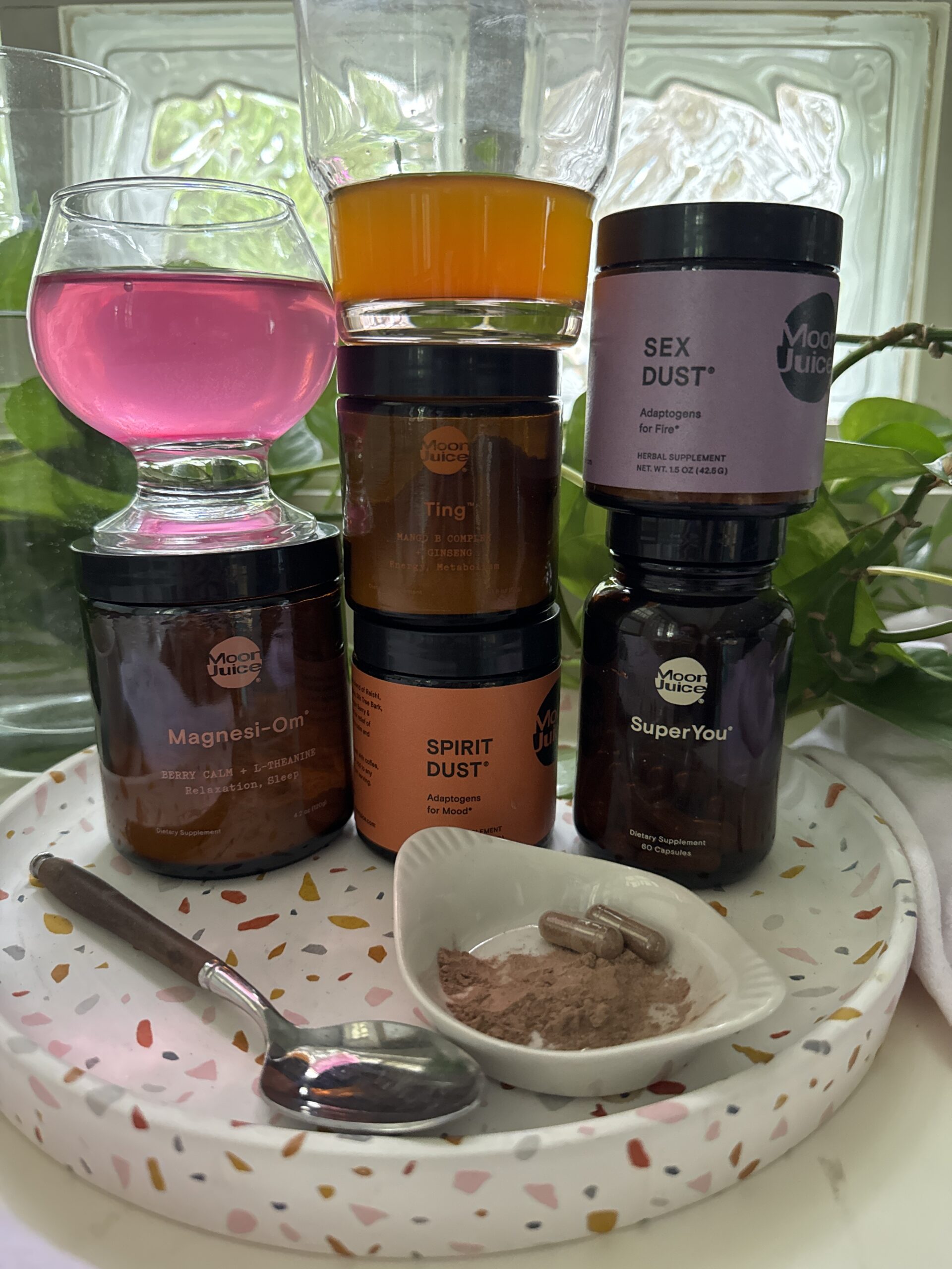 Three stacked jars labeled "magnesi-om," "spirit dust," and "sex dust," next to a dish with powder and spoon, and glasses of pink and orange liquid above.