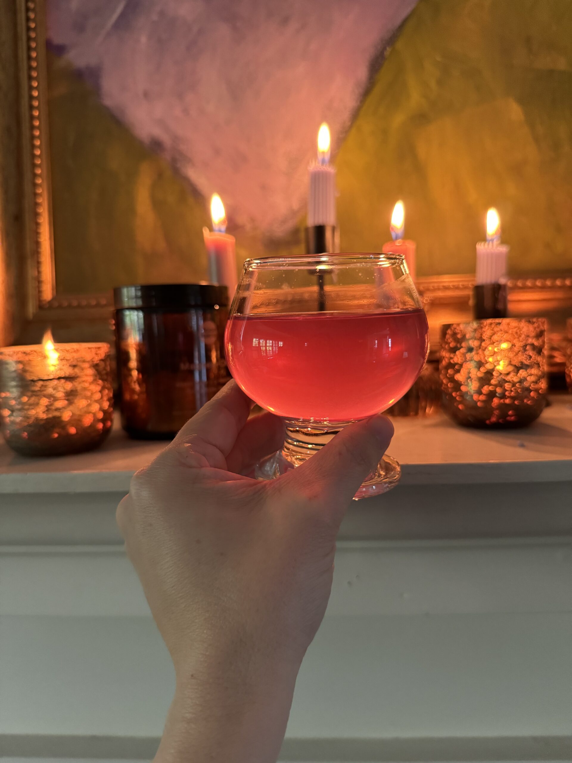 A hand holding a glass of red drink, with lit candles and decorative lights in the background, creating a warm ambiance.