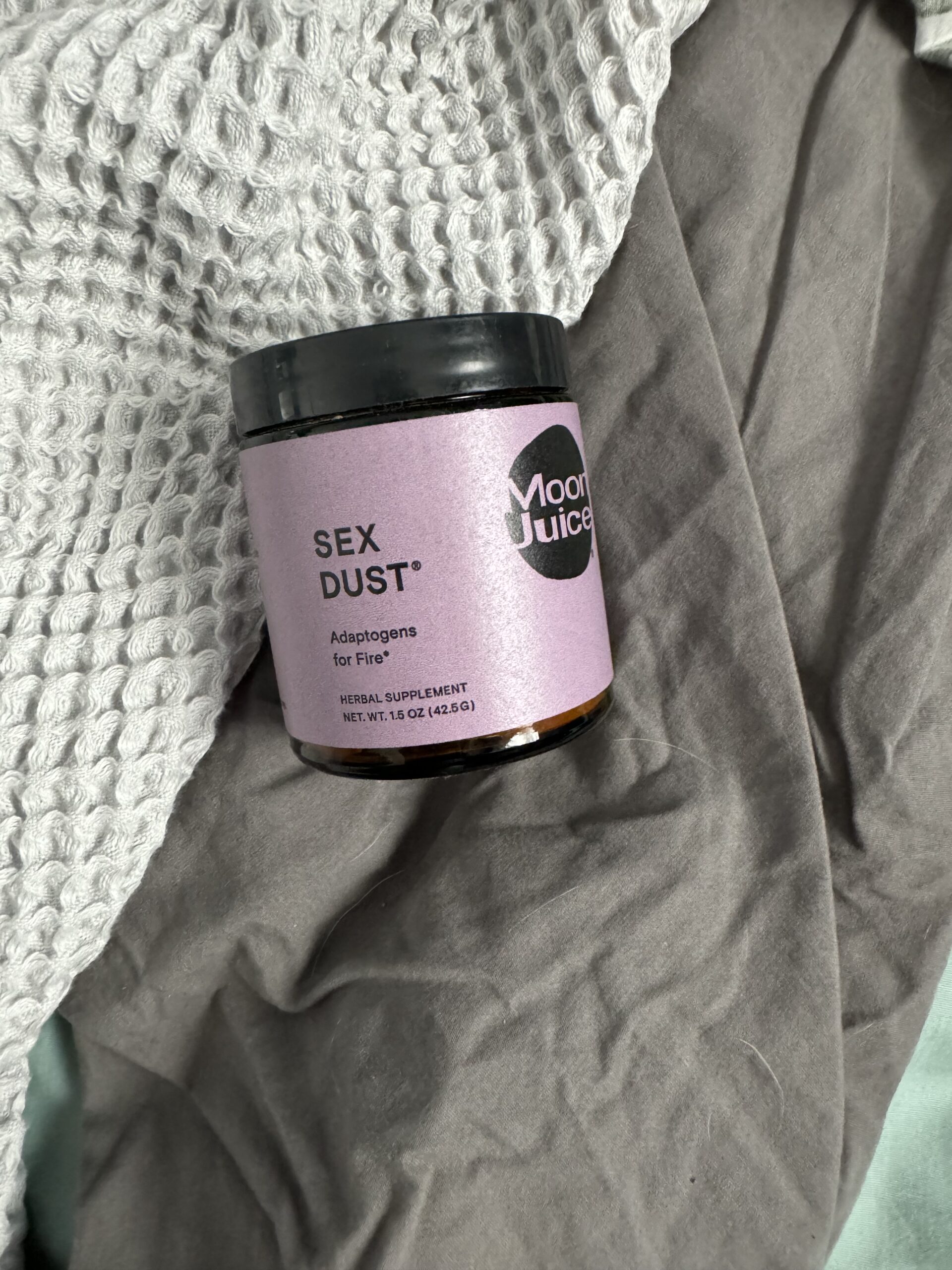 A jar of "sex dust" by moon juice, an adaptogens dietary supplement, resting on a crumpled grey fabric with a knitted blanket partially visible.