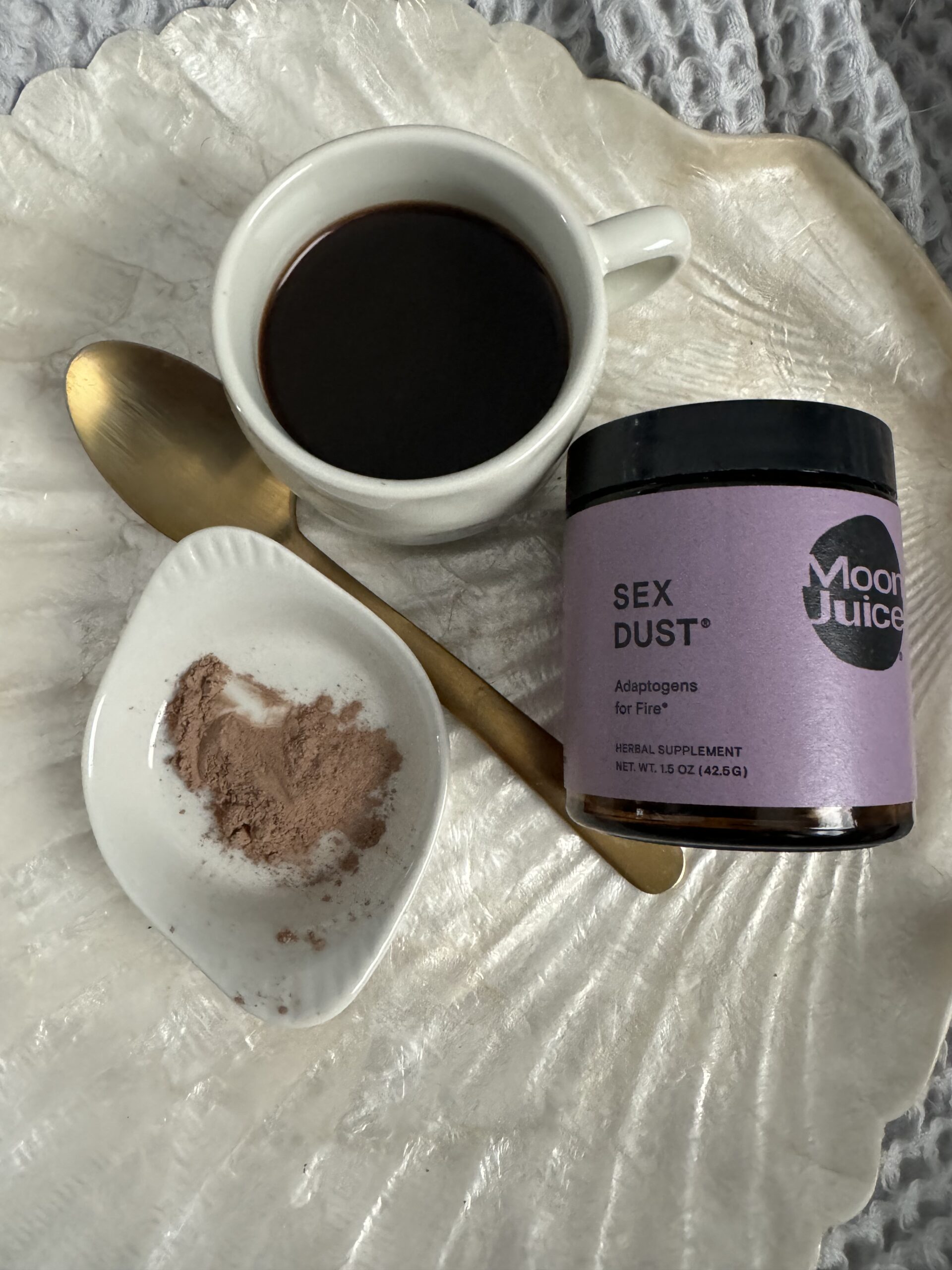 A cup of coffee with a gold spoon, next to a jar labeled "sex dust" by moon juice, and a small dish with powder on a textured white background.