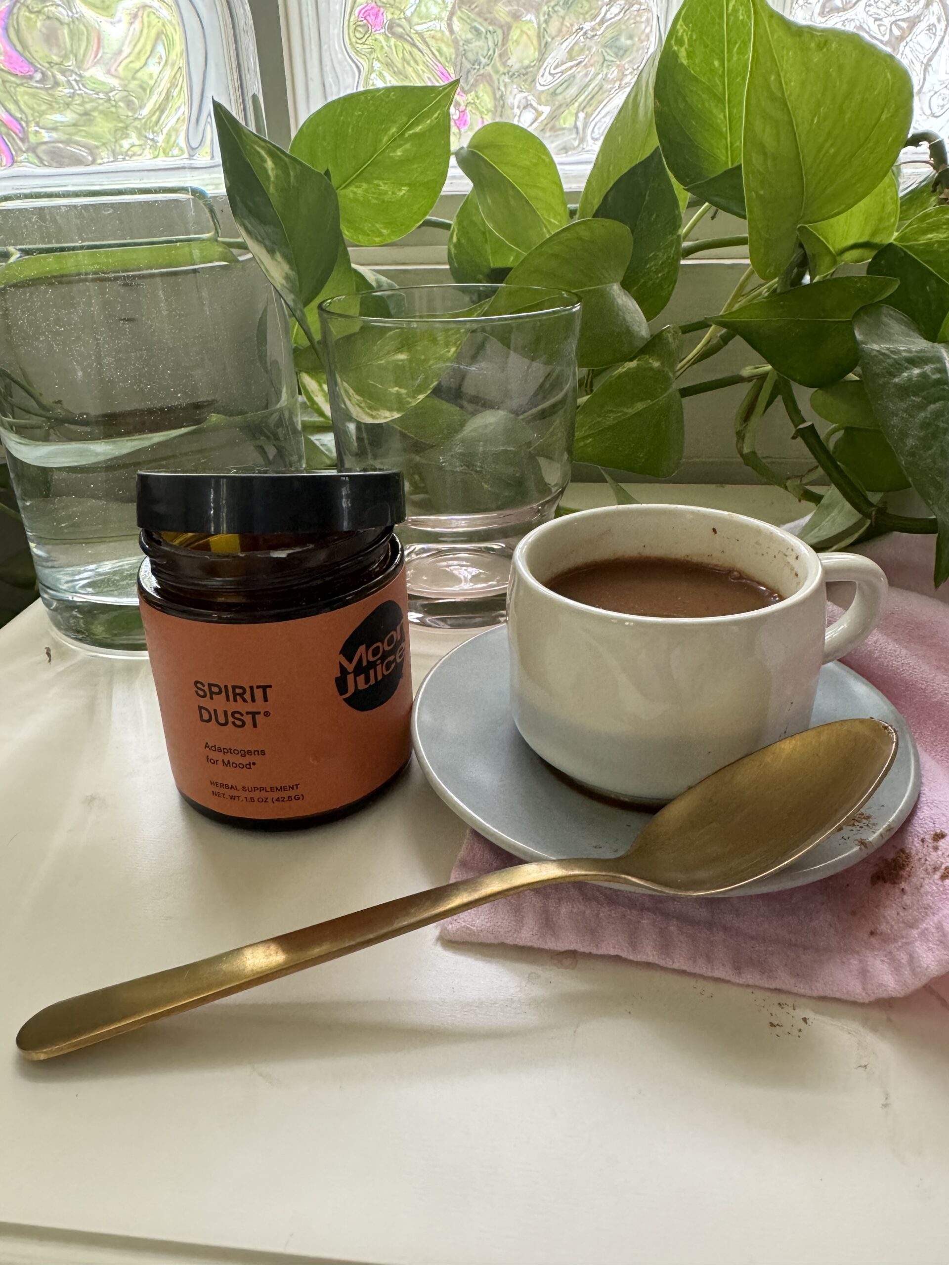 A cup of coffee on a saucer with a spoon, next to a jar labeled "spirit dust" and a glass of water, surrounded by houseplants on a table.