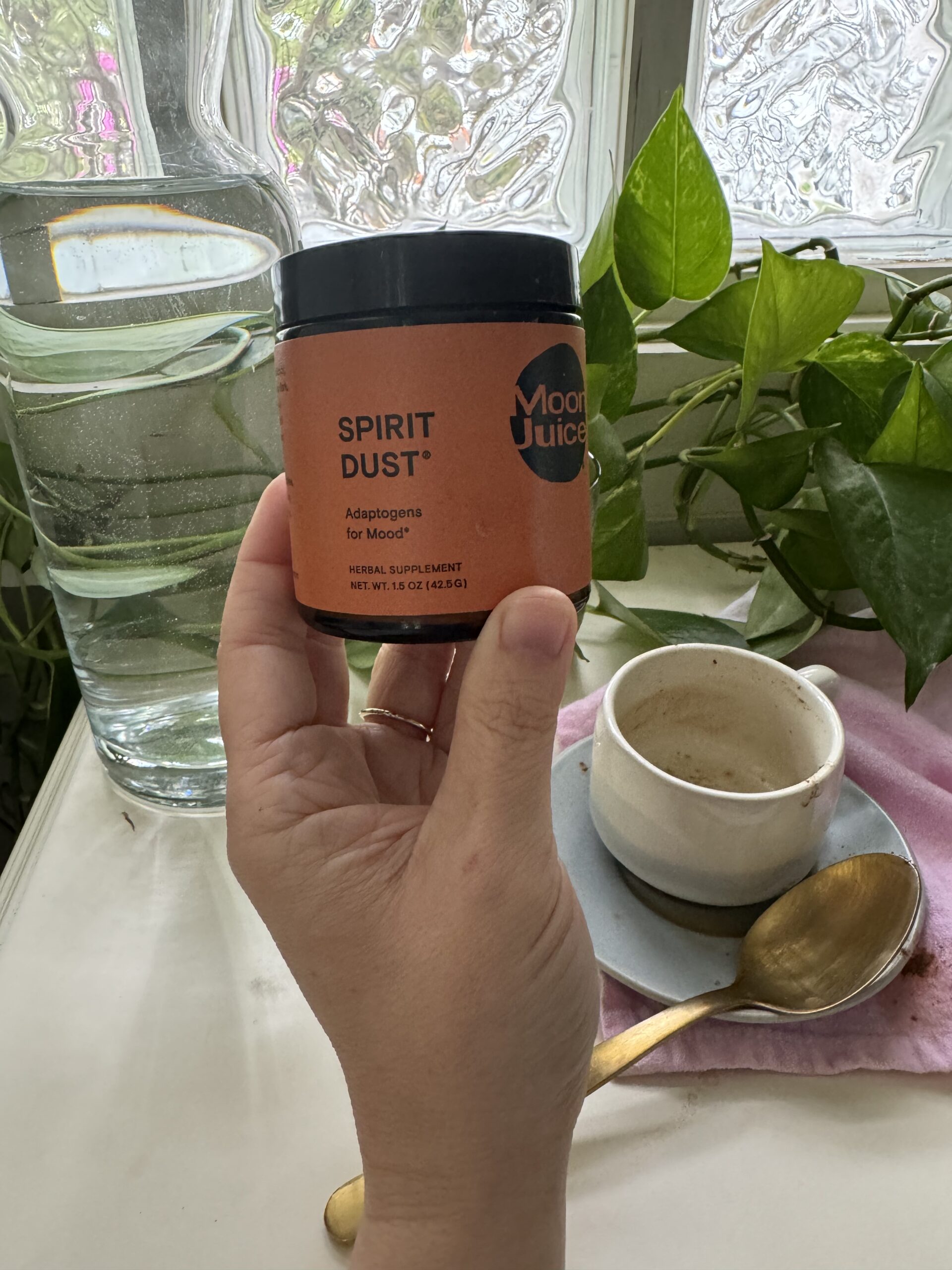 A hand holding a container of moon juice spirit dust supplement next to a cup of beverage and green plants by a window.