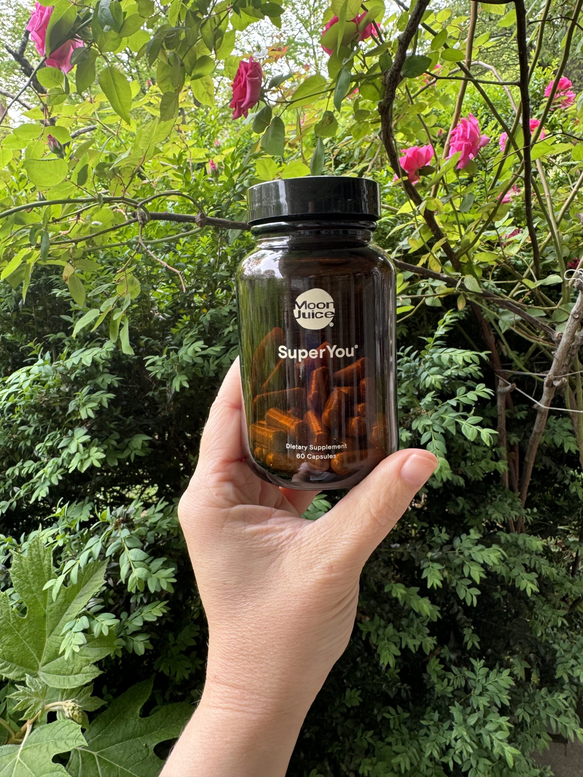 A hand holding a bottle of "moon juice superyou" dietary supplement in front of a green, leafy bush with pink flowers.