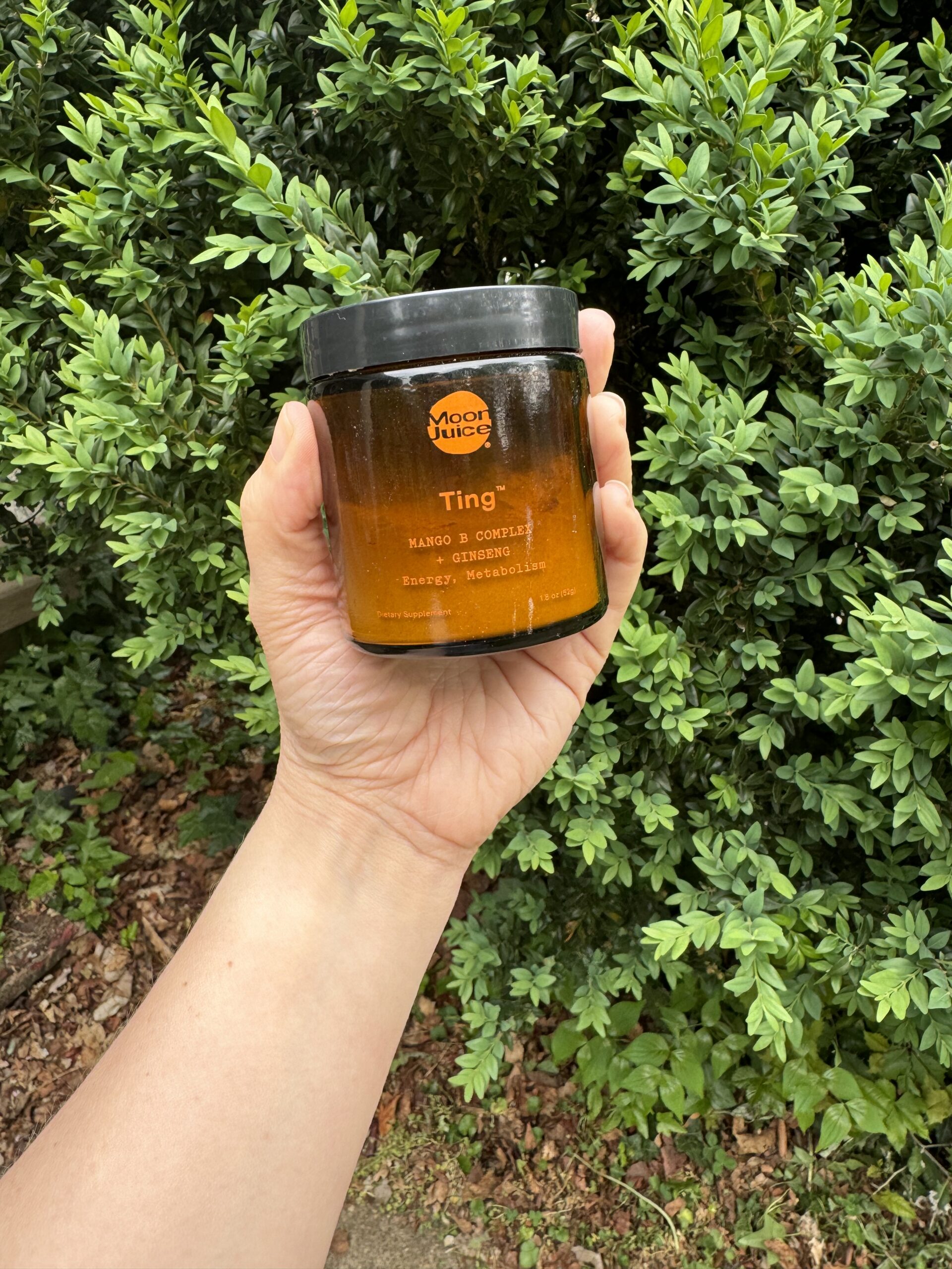 A person holds a jar of mango & coconut ting body scrub outdoors, surrounded by lush green leaves.