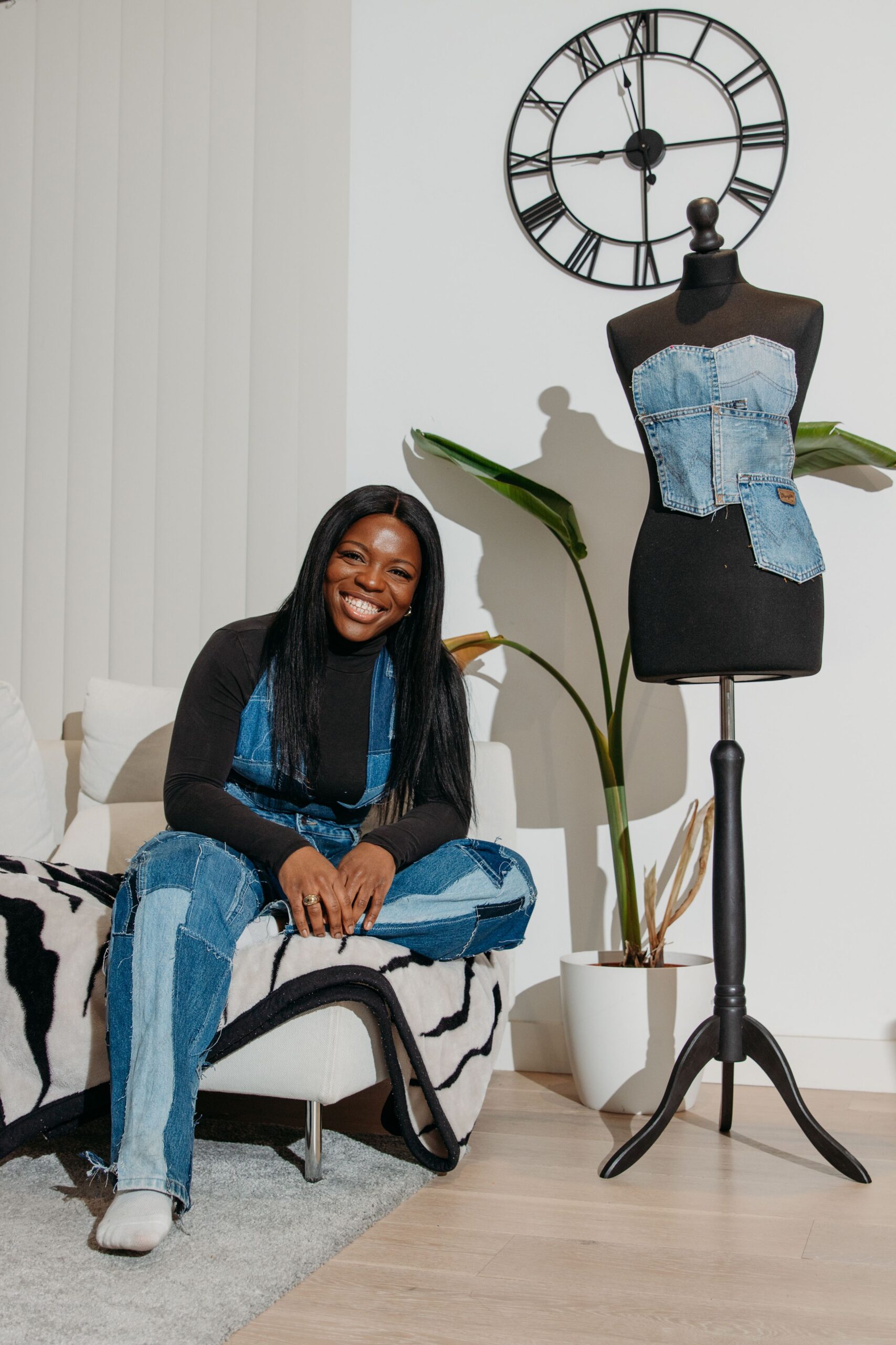 A smiling woman sits on a white sofa beside a mannequin wearing a denim jacket in a stylishly decorated room.