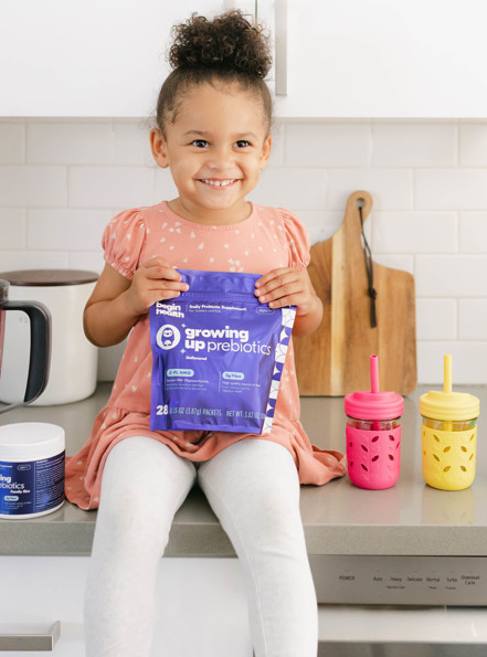 A young girl sitting on the counter next to colored water bottles, holding a packet of Begin Health's growing up prebiotics.
