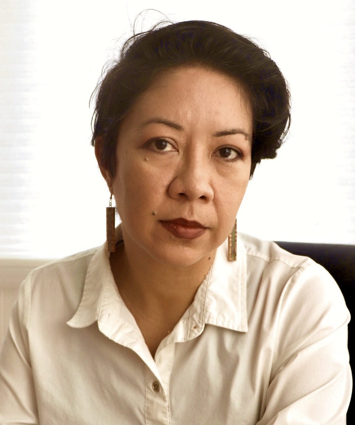 A middle-aged woman with short dark hair wearing a white blouse and triangular earrings, sitting against a lightly shaded background, facing the camera with a serious expression.