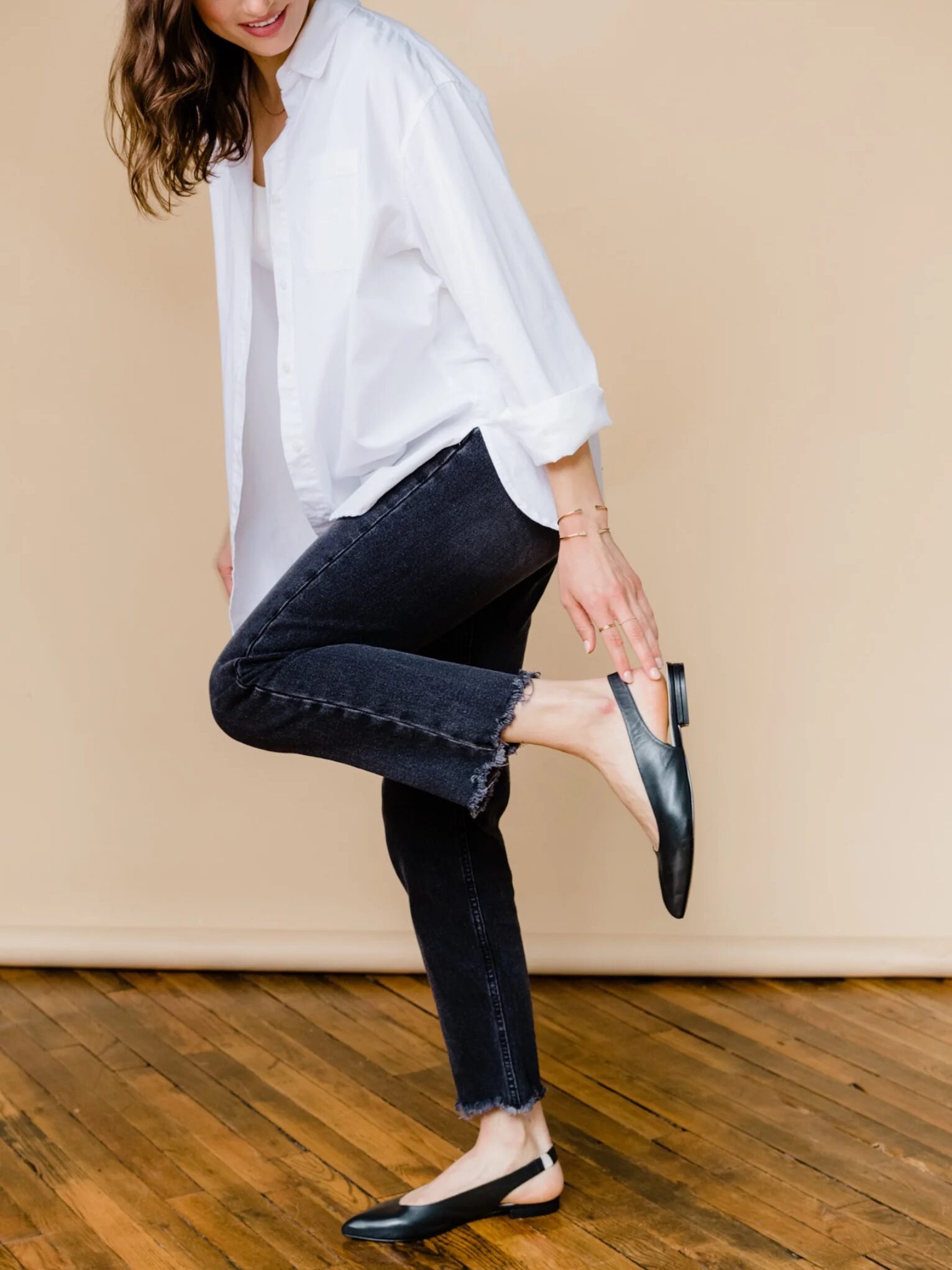 Woman in a white shirt and distressed jeans adjusting her black flat shoe, standing against a beige background.