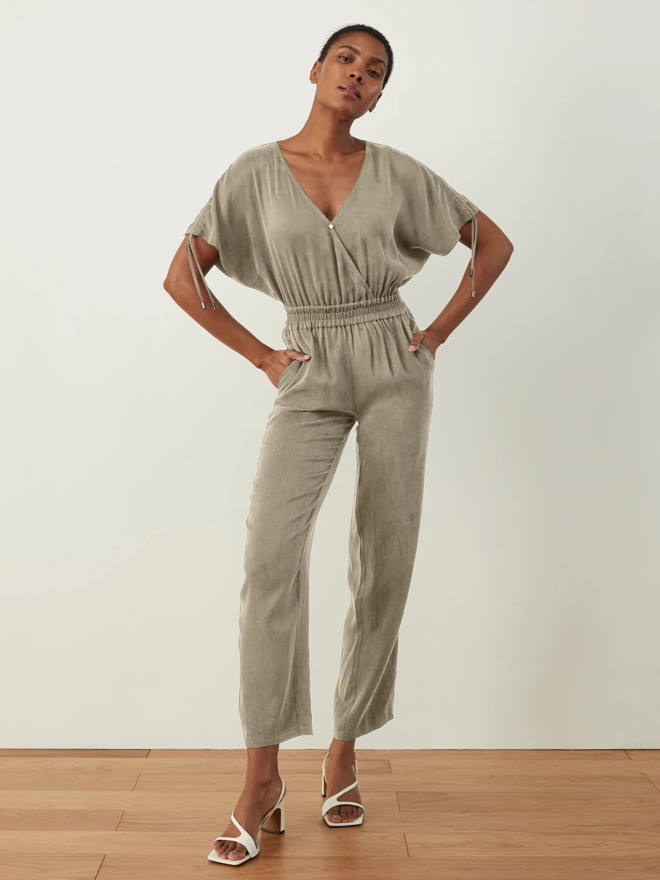 A woman stands confidently with her hands on her hips, wearing a grey jumpsuit and white heels, against a neutral background.