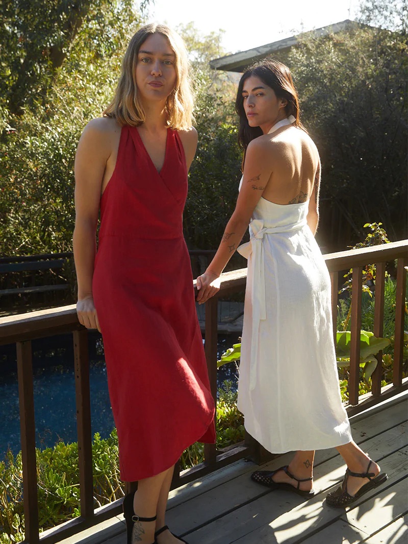 Two women in elegant dresses standing on a wooden deck, holding hands and looking over their shoulders at the camera.