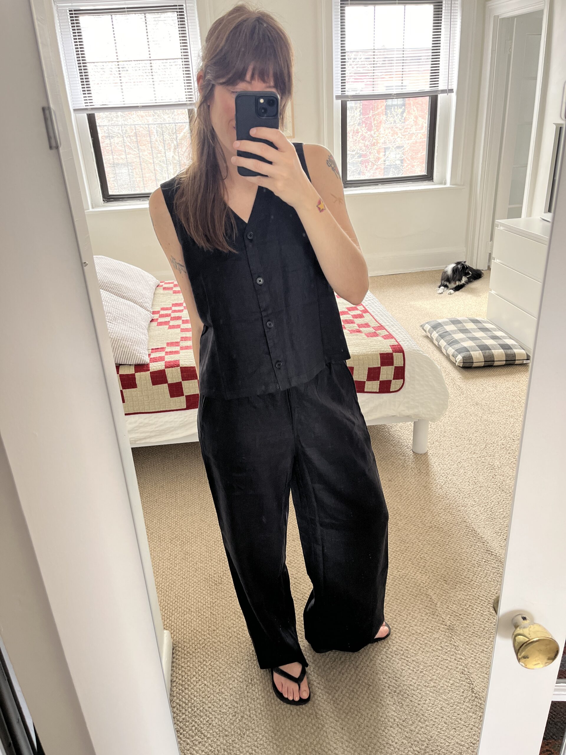 Woman in a black sleeveless jumpsuit taking a mirror selfie in a bedroom, with a dog lying on a bed in the background.