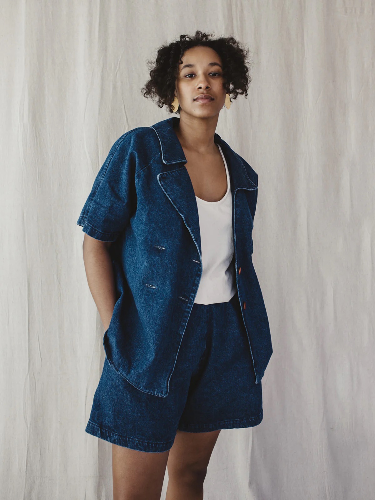 A person wearing a denim jacket and shorts set, standing against a textured beige background.