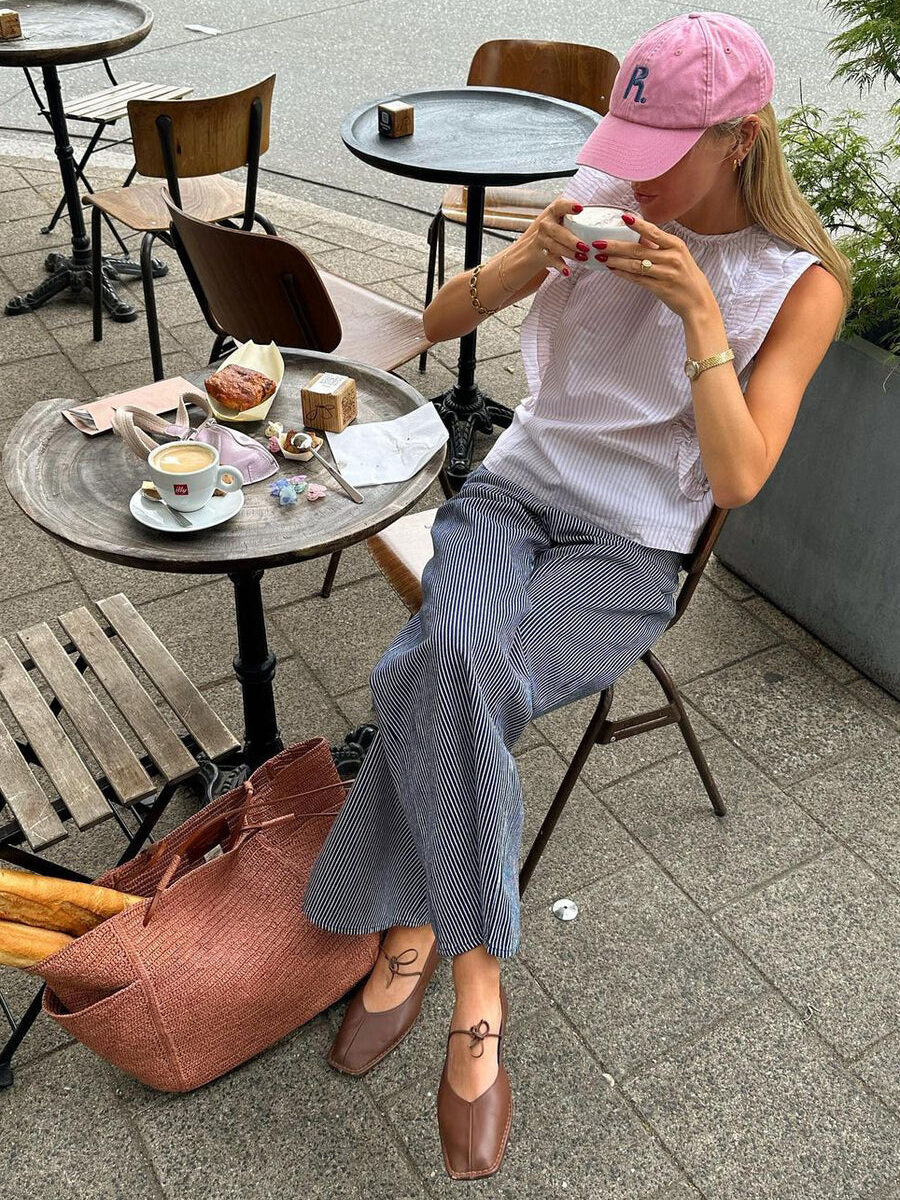 A woman in a pink hat and blue striped pants sits at a café table drinking coffee, with a pastry and a book on the table.