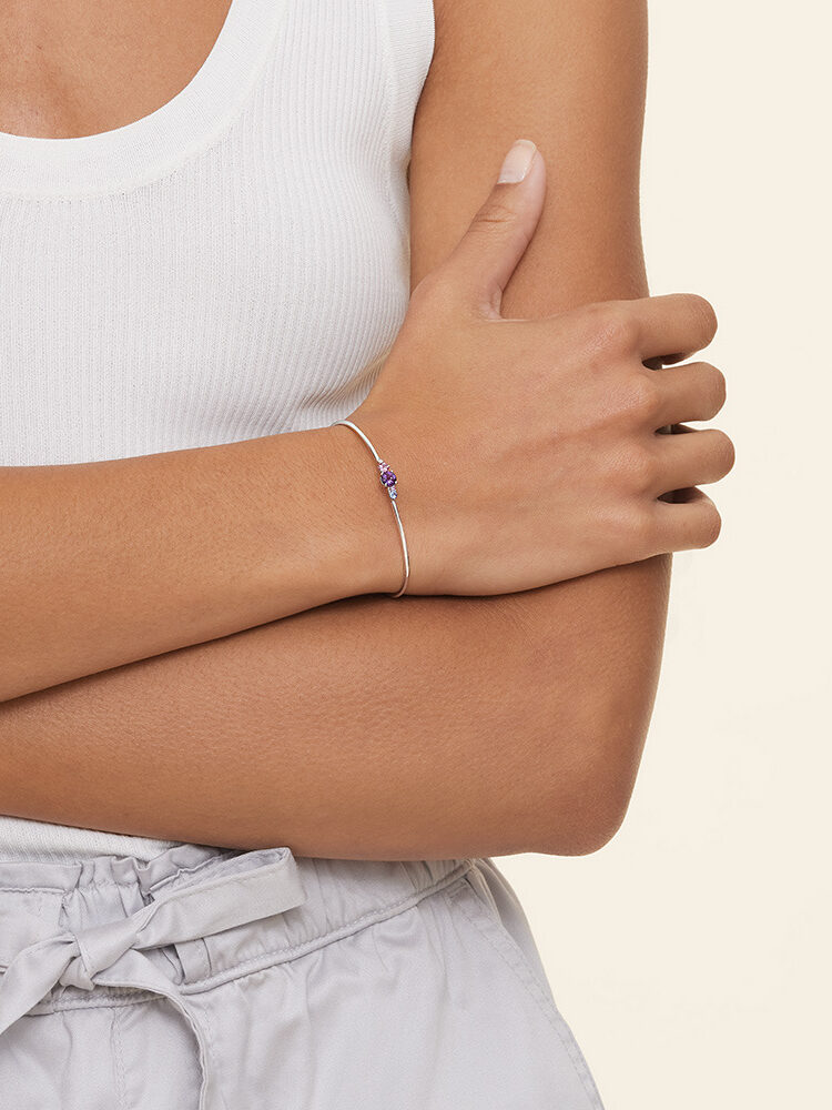 Woman in white tank top wearing a delicate bracelet with a blue gemstone, her arm crossed over her torso.