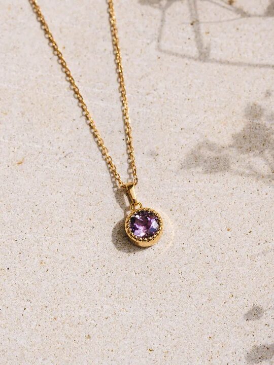 A pendant with a purple gemstone on a gold chain, presented on a sandy surface with a shadow of foliage.