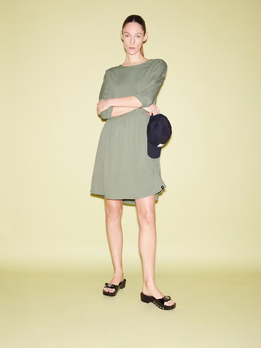 Woman in a green dress standing against a yellow background, holding a navy cap, with her arms crossed.