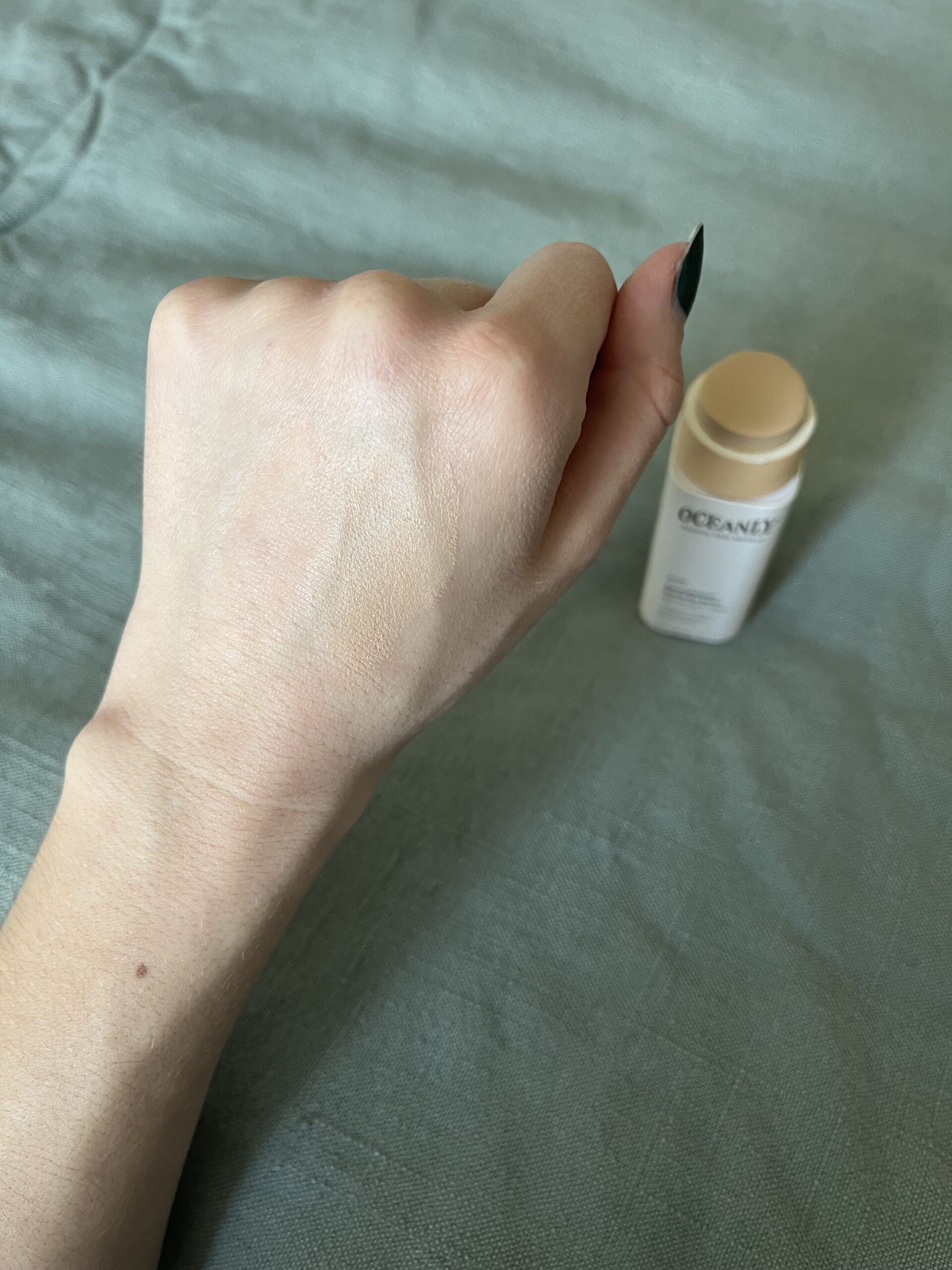A person's hand showing a swatch of light beige foundation next to a small bottle of oceane makeup on a grey fabric surface.