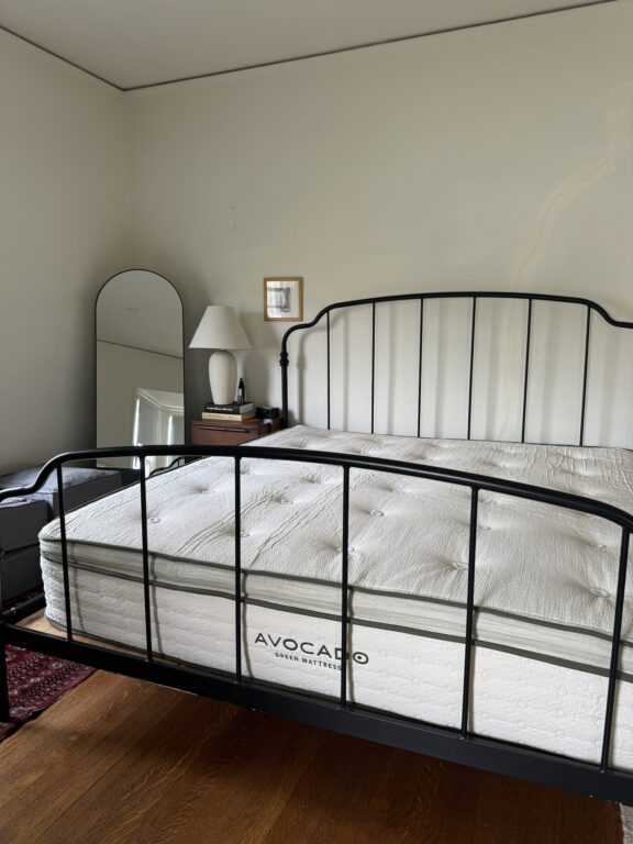 A tidy bedroom featuring a black metal frame bed with a white puffy mattress, matching bedside table with lamp, and an arched mirror against a light gray wall.