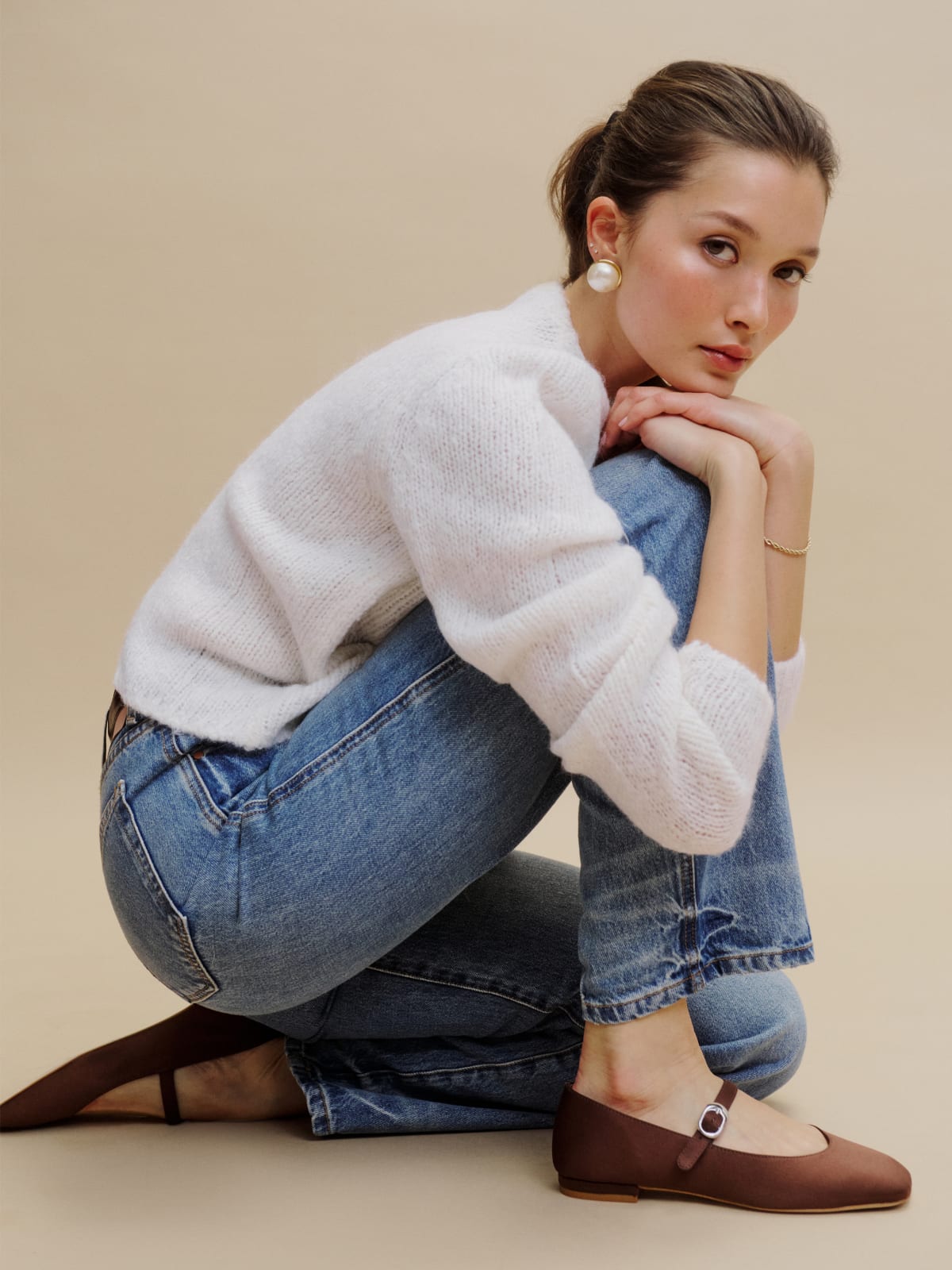 A young woman in blue jeans and a white sweater crouches, wearing brown flats and large pearl earrings, looking at the camera with a neutral expression.