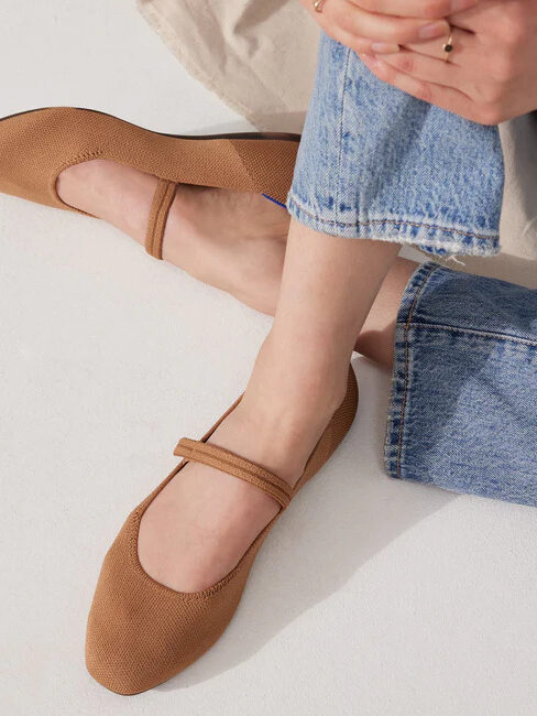 A person seated wearing brown mary jane style flats, paired with denim jeans, fingertips resting on one ankle.