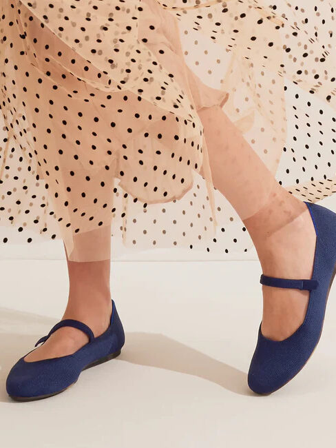 Close-up of a person's lower legs wearing blue mary jane flats and a beige polka dot skirt against a pale background.
