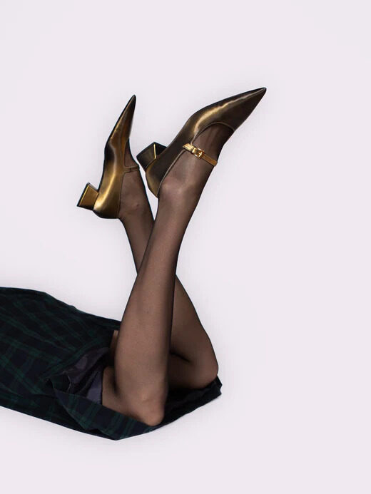 A person wearing gold high heels and black pantyhose with their legs up against a white background.