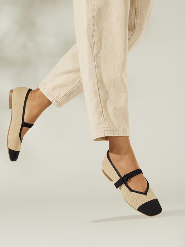 A woman's feet mid-air in beige pants and black-beige strapped flat shoes, with a soft, neutral background.
