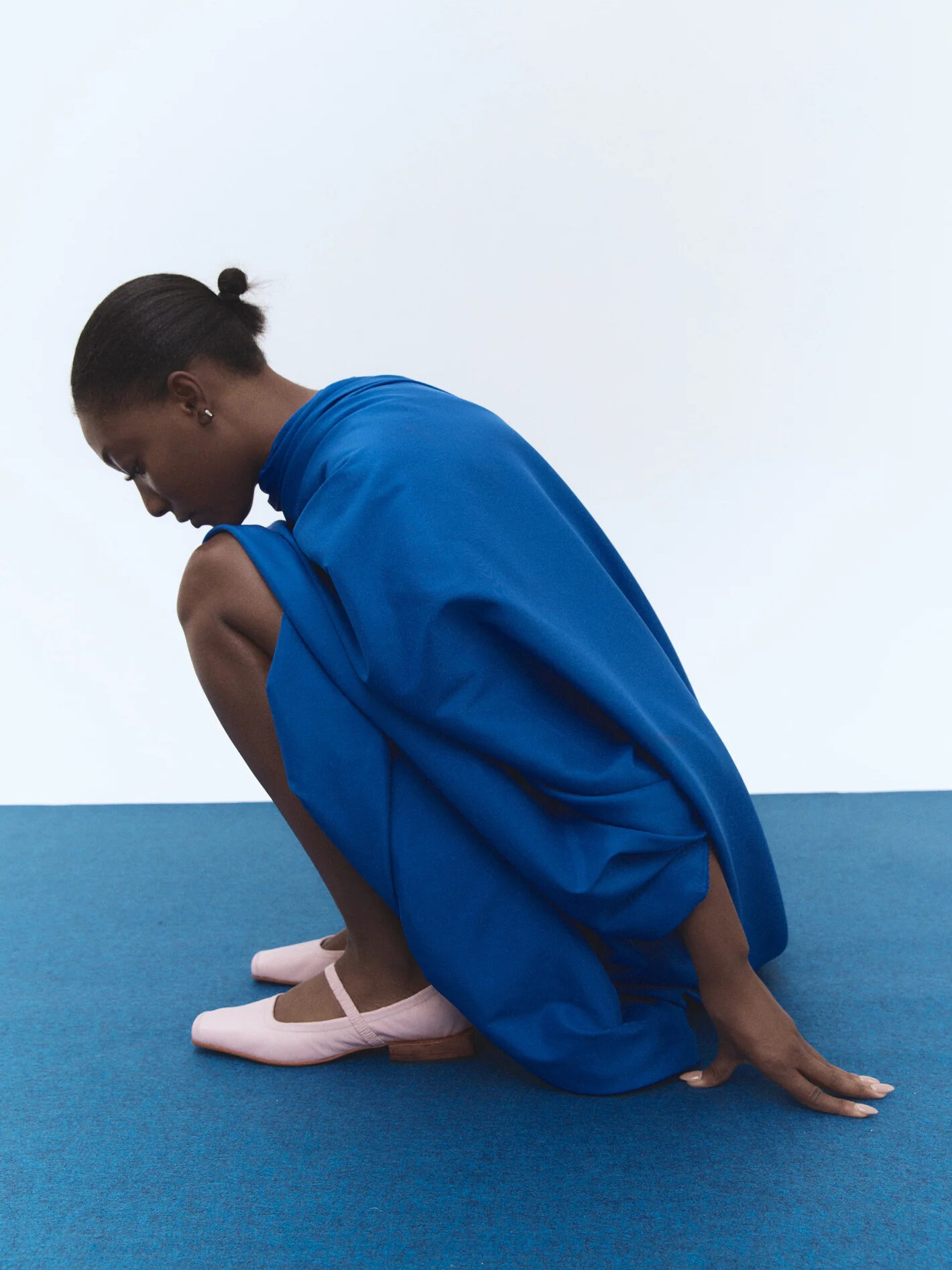 A woman in a flowing blue dress and pink shoes crouches on a blue mat against a white background.