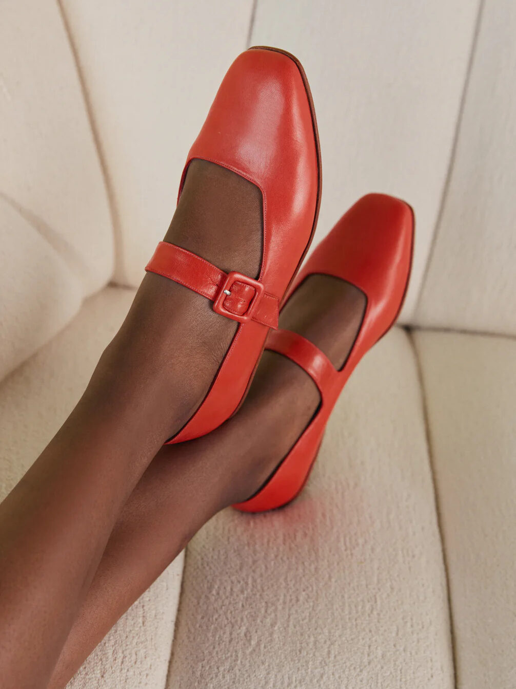 A person wearing bright red mary jane shoes with buckled straps, positioned with feet crossed, against a light textured background.