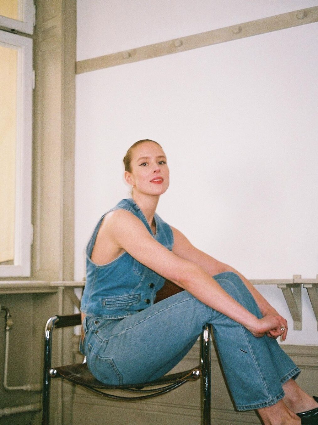A model in a denim vest and jeans, seated on a chair with their legs over the armrests. 