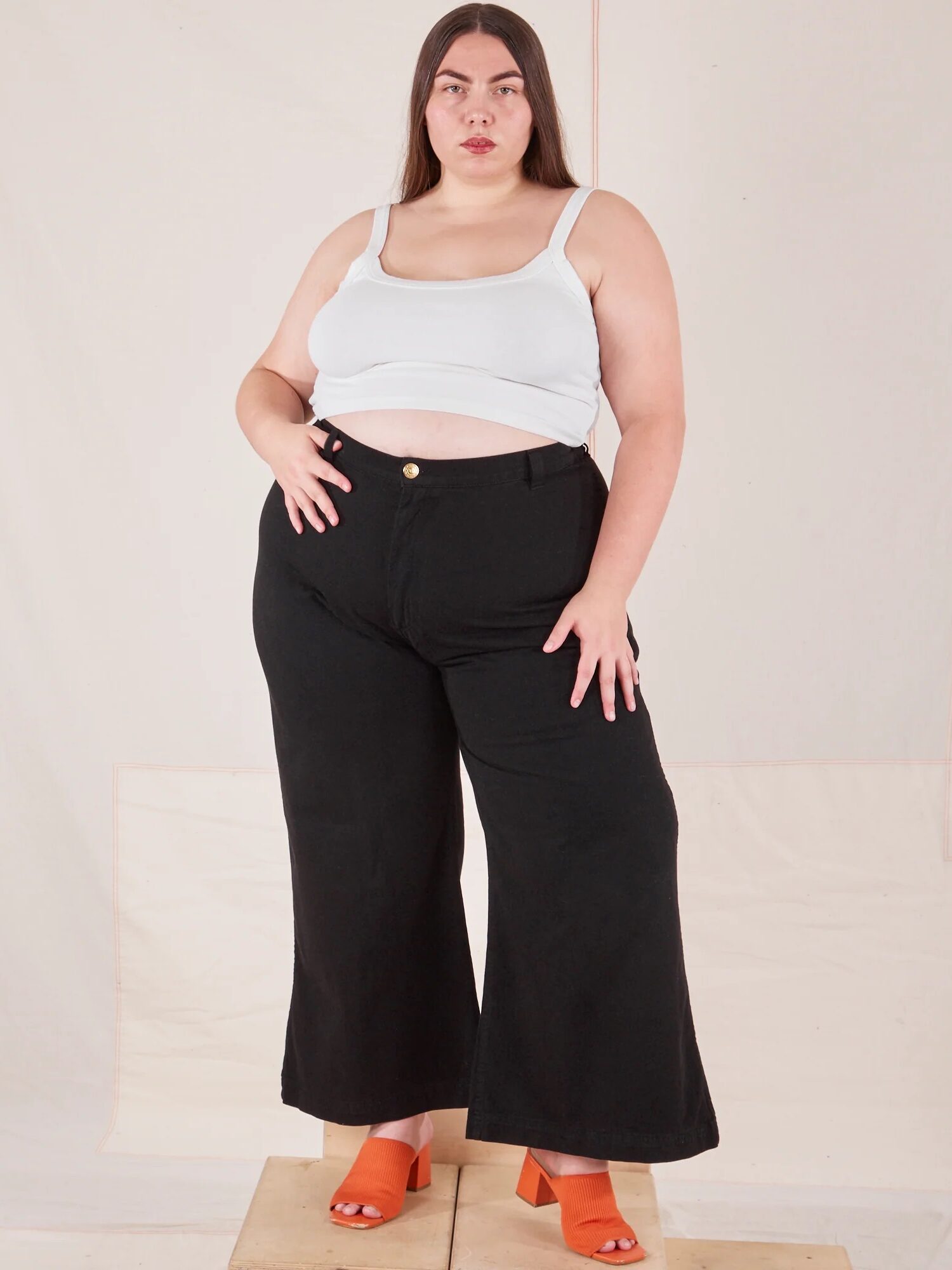 A woman in a white crop top and black palazzo pants stands confidently, with one hand on her hip.