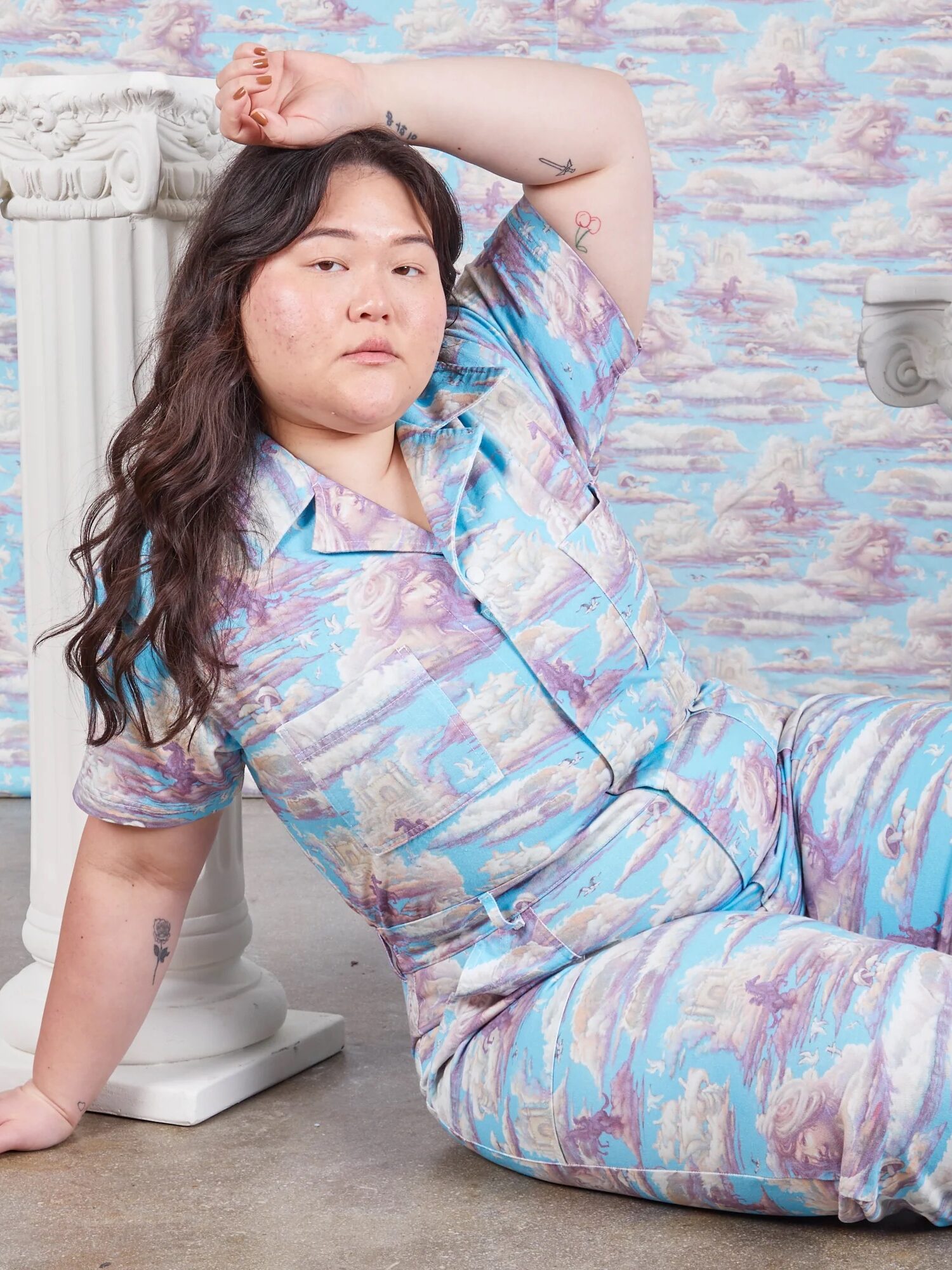 A woman in a matching pastel landscape-print pajama set posing seated with one arm raised, against a similarly patterned backdrop.