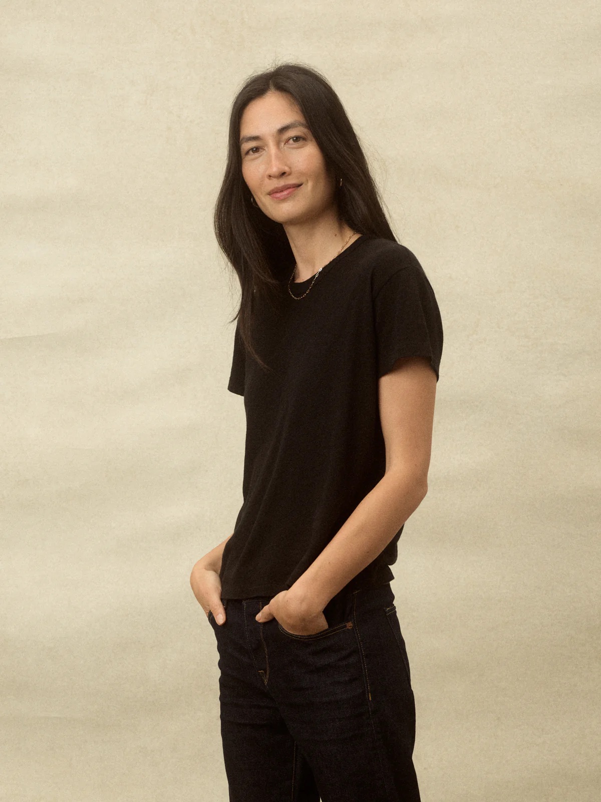 A woman in a black t-shirt and jeans stands casually with one hand in her pocket against a beige background.