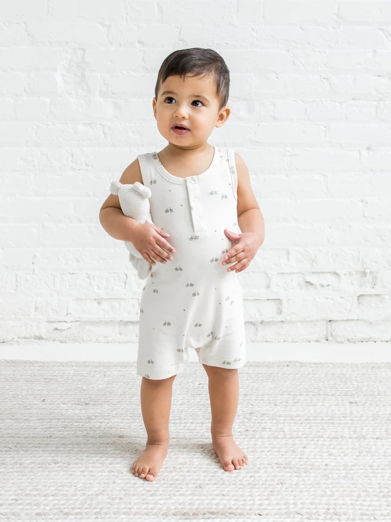 A toddler in a onesie stands on a white rug, holding a plush toy, with a brick wall in the background.