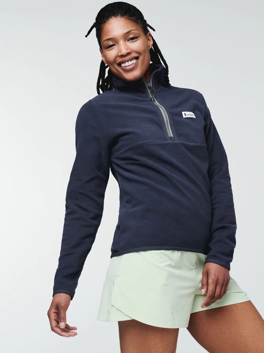 A woman smiles at the camera, wearing a dark blue pullover and light green shorts. she has braided hair and stands against a light gray background.