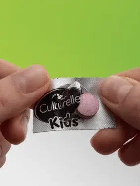 Two hands holding a blister pack of culturelle kids chewable probiotic tablets against a green background.