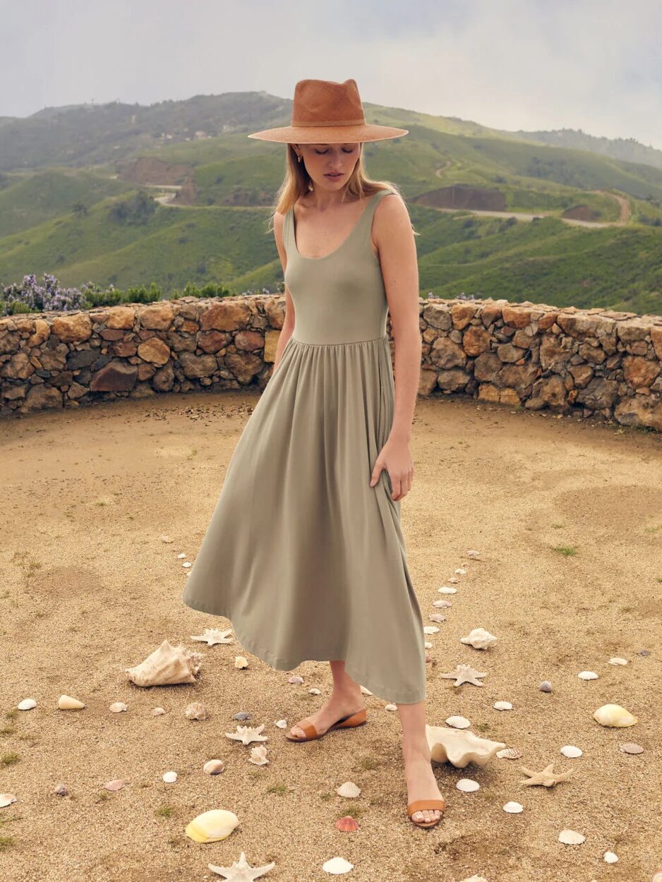 A woman in a green dress and a wide-brimmed hat walks pensively among scattered white seashells on a dirt ground, with a hilly landscape in the background.