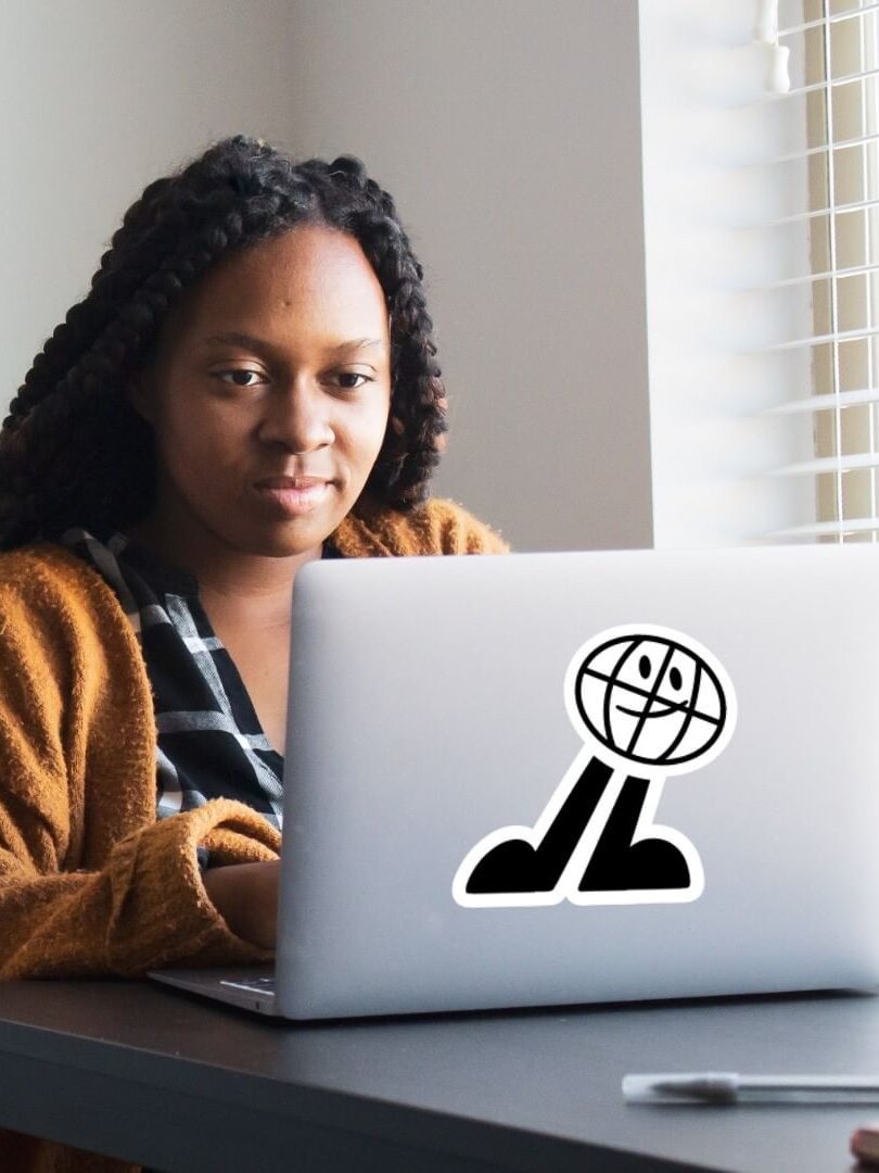 A woman with braided hair, wearing a beige cardigan, works at a laptop with a globe sticker, in an office setting by a window.