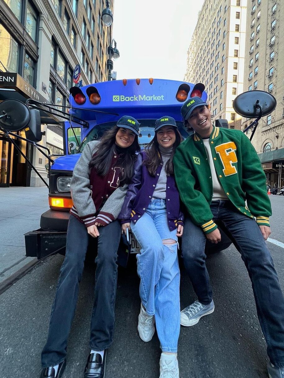 Three young adults smiling, seated on a blue scooter adorned with balloons, in a city street setting.