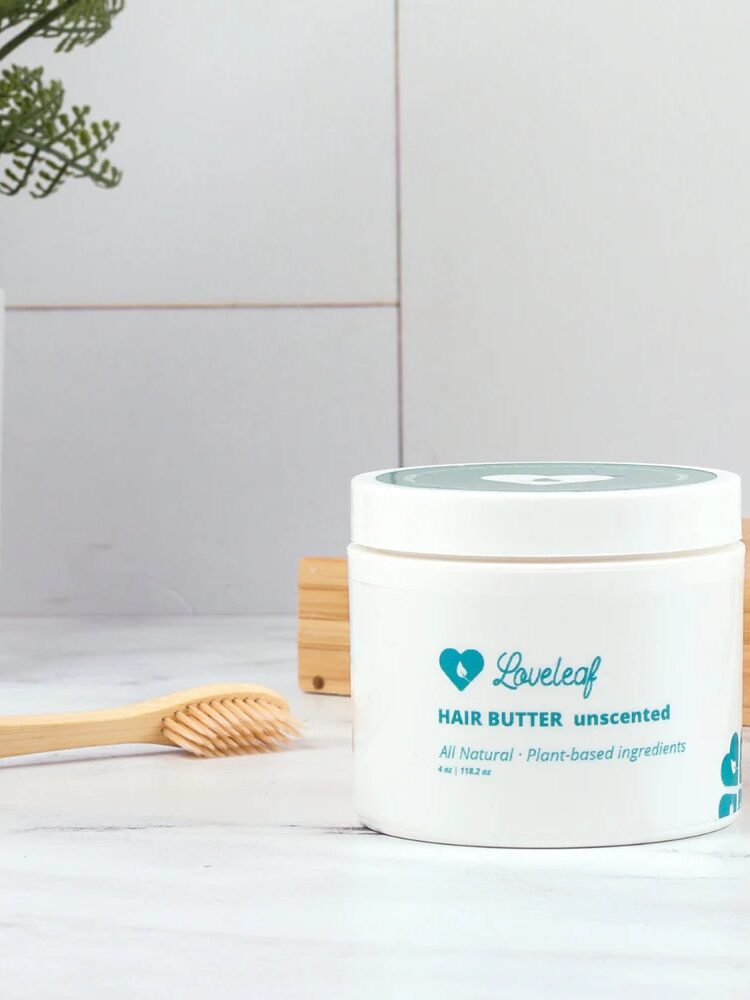 A jar of unscented loveleaf hair butter with a bamboo toothbrush on a wooden tray beside it, placed against a white tile background.